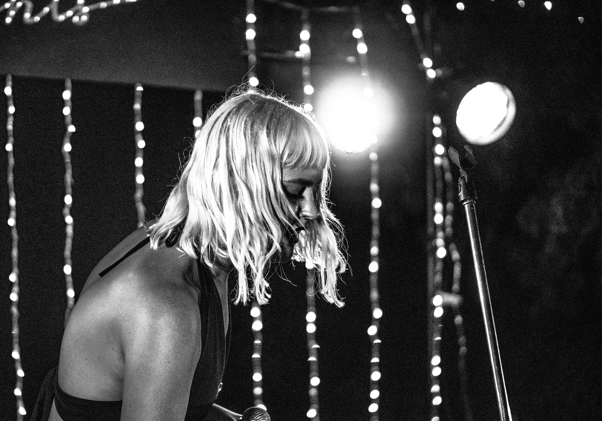 A woman with long blonde hair and glasses, singing into a microphone on stage, illuminated by bright stage lights with a dark background and decorative lights.