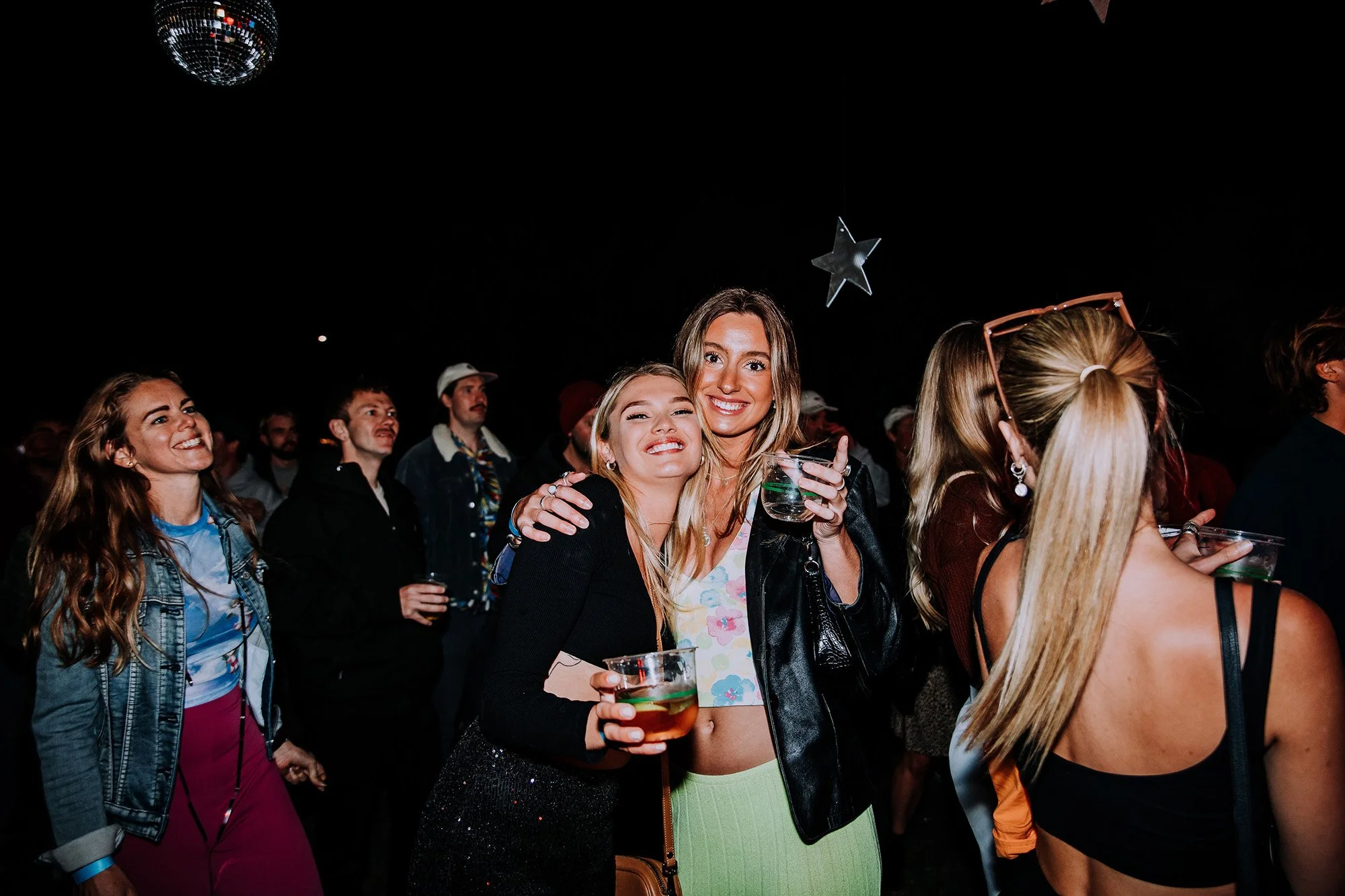 A group of young people at a party or concert, some holding drinks, enjoying themselves under a dark sky with disco balls hanging above.