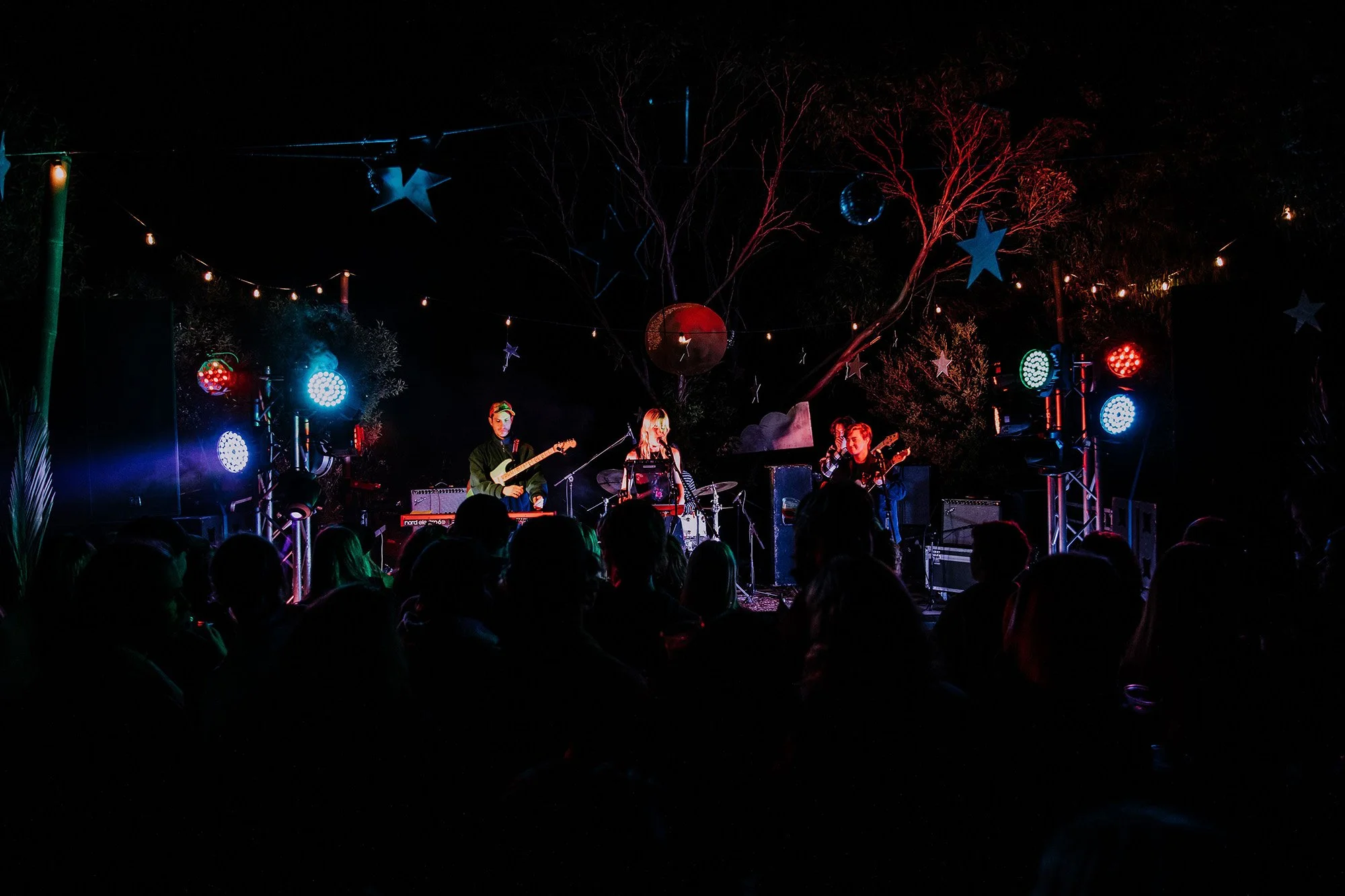 Nighttime outdoor concert with band performing on stage, colorful lights, stars and disco ball hanging from trees, audience in silhouette, festive atmosphere.