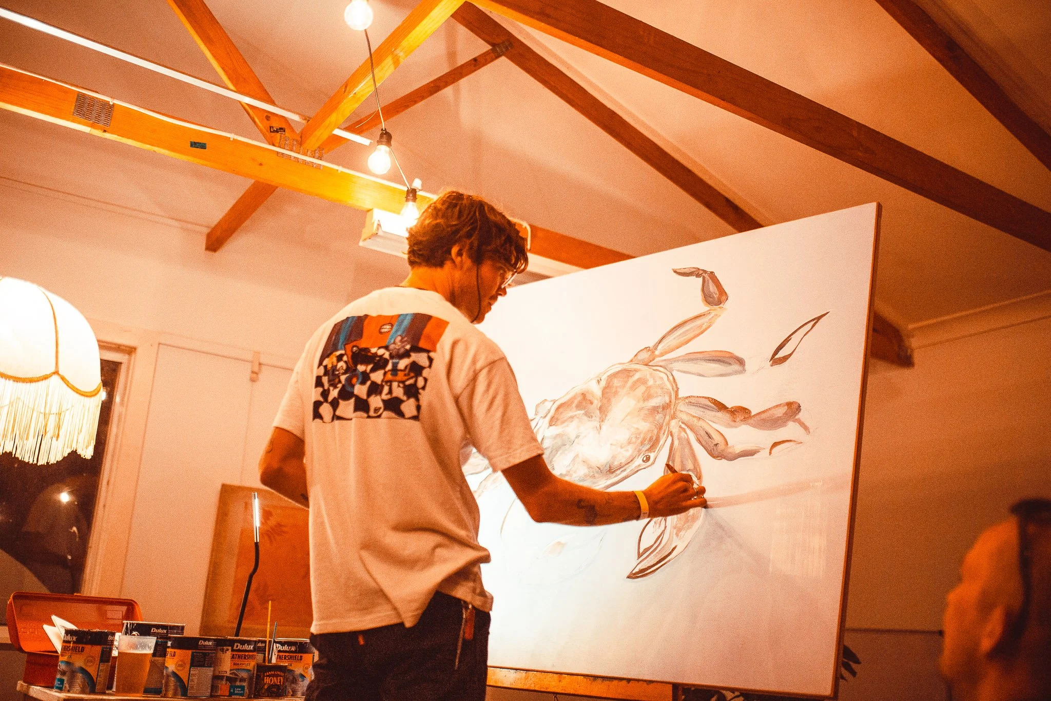 A man with glasses and a tattoo on his right arm, wearing a colorful patterned shirt, is painting a large canvas featuring a rabbit and flowers in a warmly lit room with wooden beams and hanging lights.