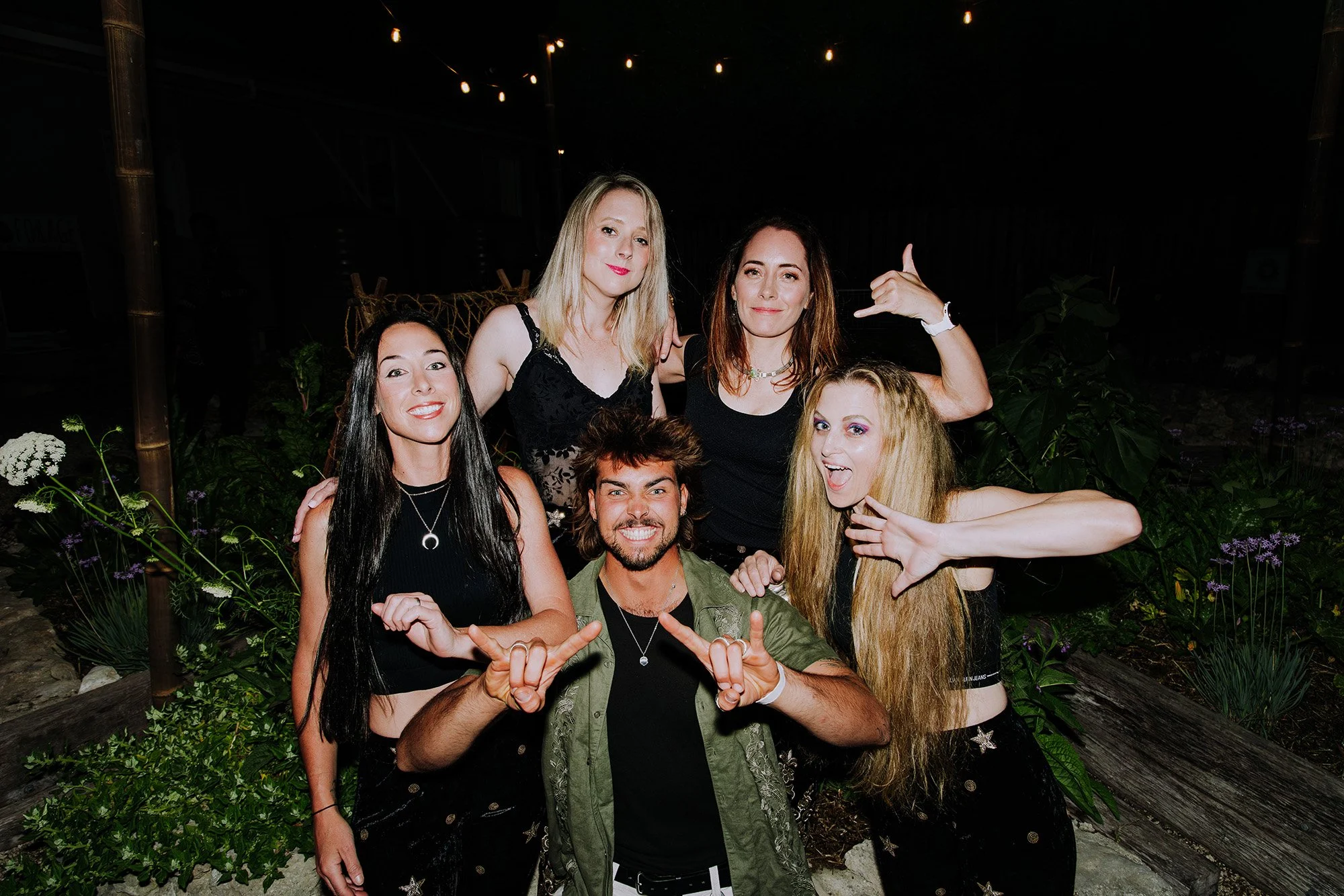 Group of six young adults smiling and posing for a photo outdoors at night, with plants and string lights in the background.