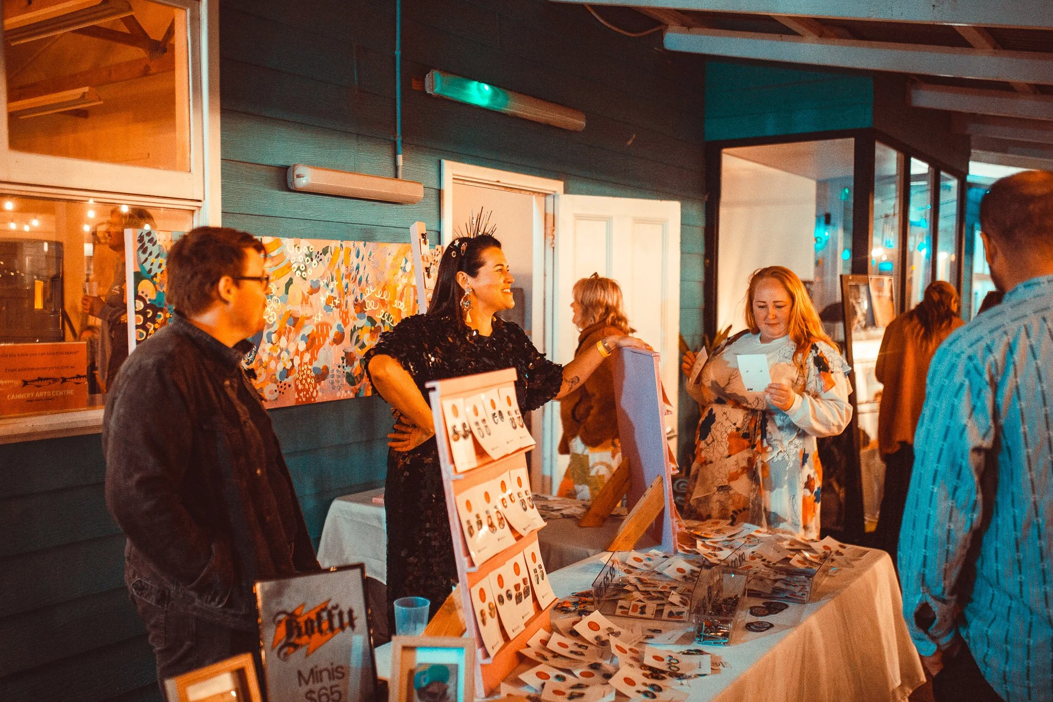 People gathered at an outdoor market stall during evening with jewelry on display and a woman smiling behind the stall.