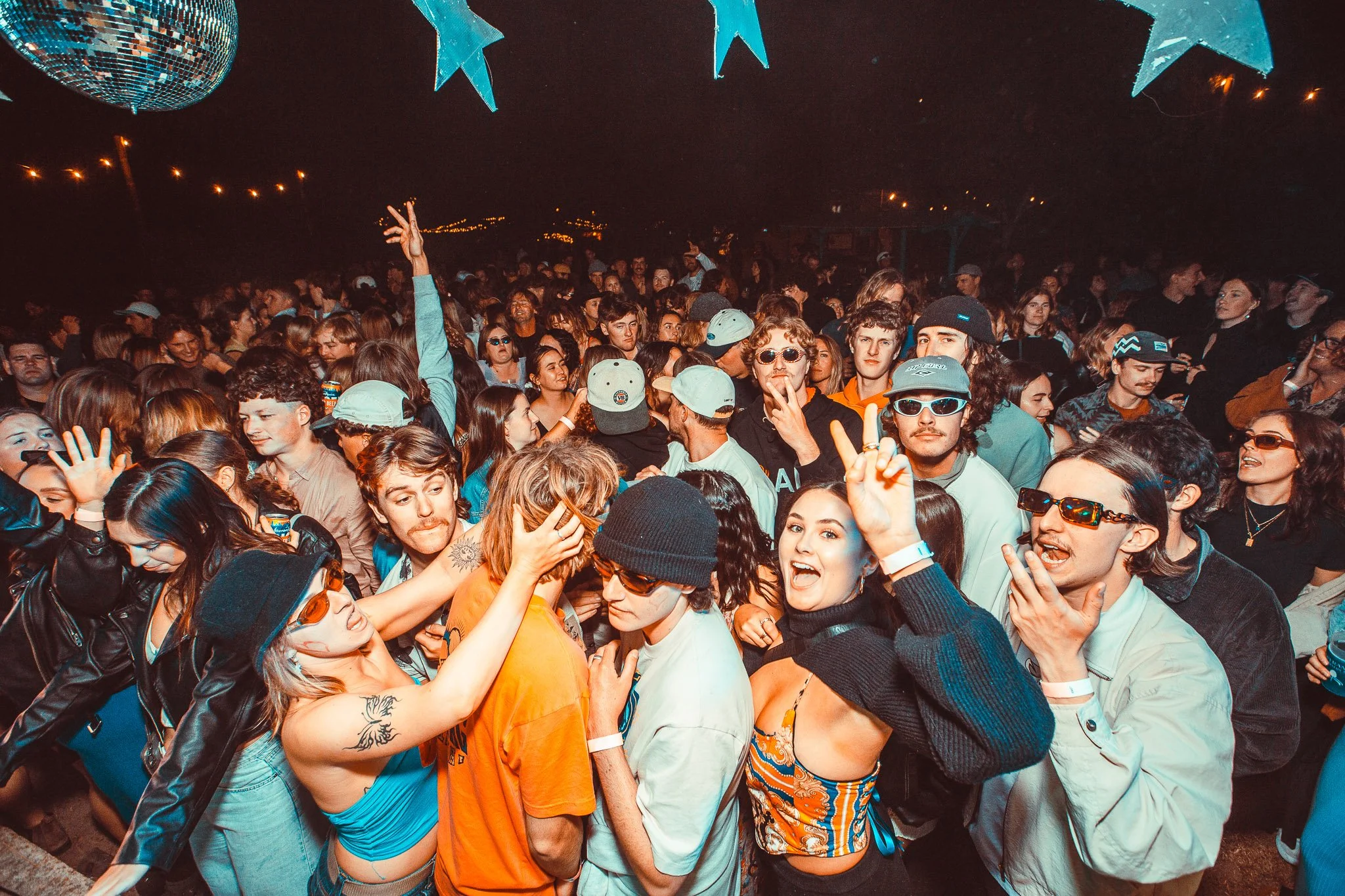 Crowd of young people at a nighttime outdoor party or music event, with some wearing sunglasses and caps, and colorful decorations hanging above.