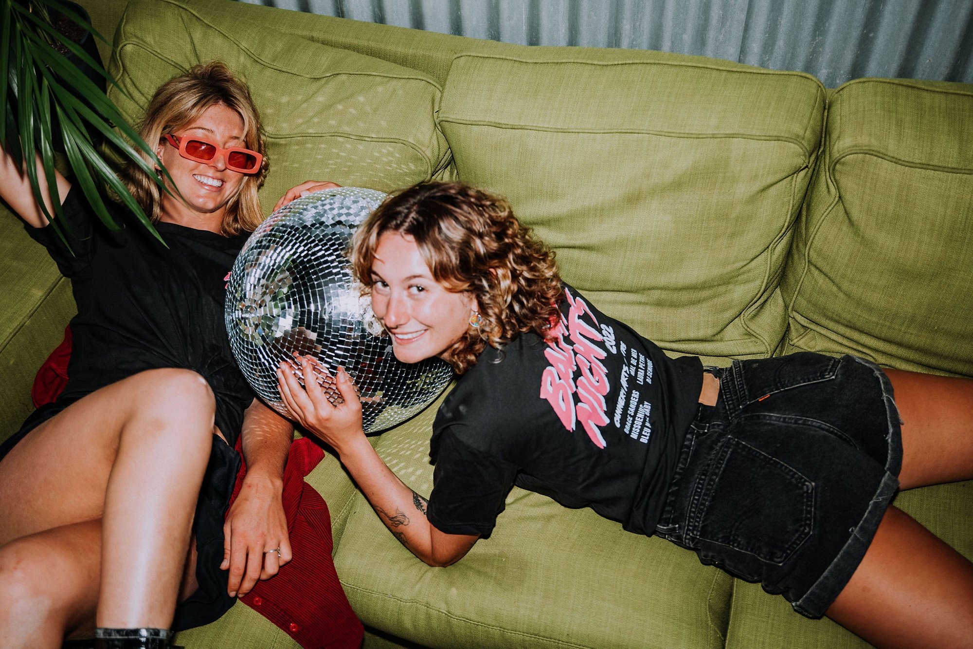 Two women lying on a green couch, holding a disco ball, smiling, one wearing red sunglasses, the other with curly hair and a graphic t-shirt.