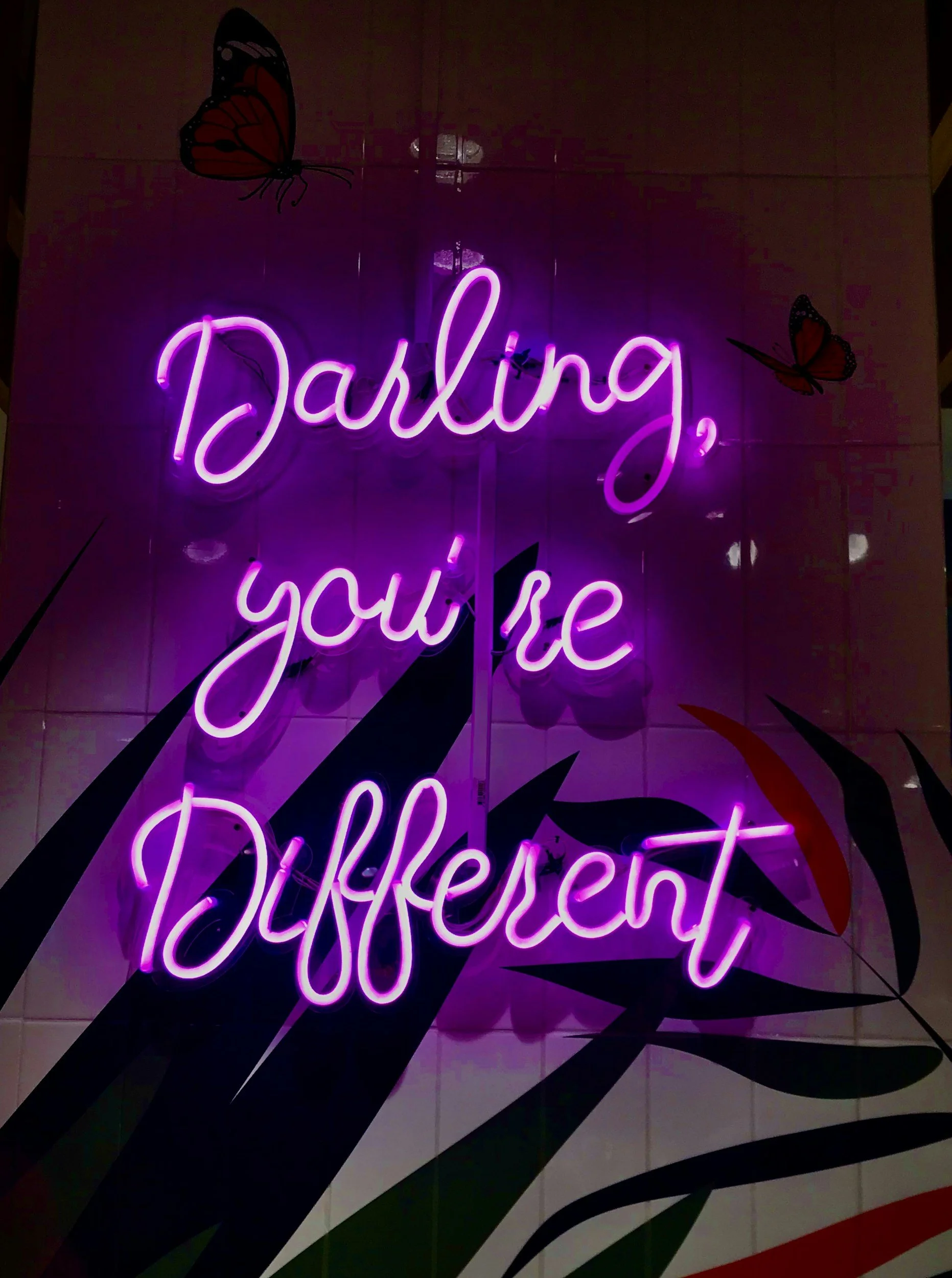 Neon sign with pink cursive text reading 'Darling, you're Dazzling' amid decorative butterflies and colorful leaf patterns.