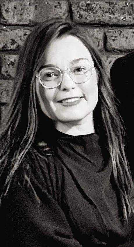 A woman with long hair and glasses smiling, sitting in front of a brick wall.