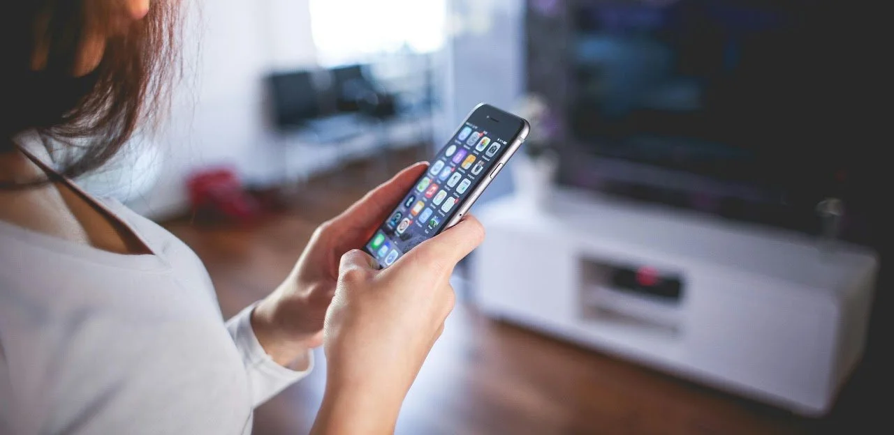 Person hält ein Smartphone mit vielen Apps auf dem Bildschirm in der Hand in einem Wohnzimmer.