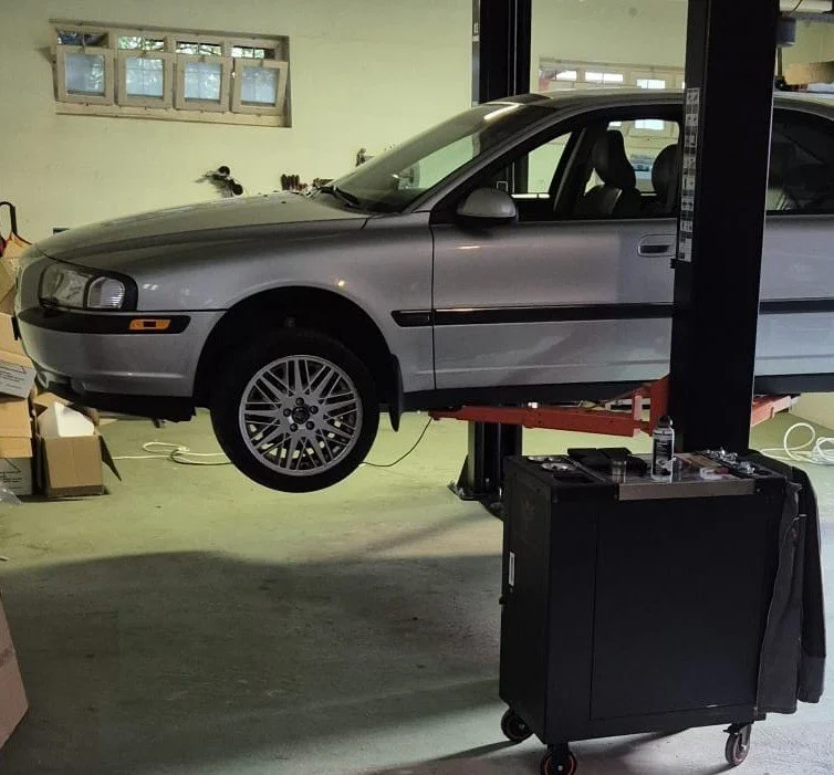 Ein silberner BMW-Fahrzeug auf einer Hebebühne in einer Autowerkstatt, ohne Räder. Im Vordergrund ein Werkzeugwagen mit Werkzeugen, neben dem Fahrzeug befindet sich eine schwarze Falltür mit einem Wasserflasche.