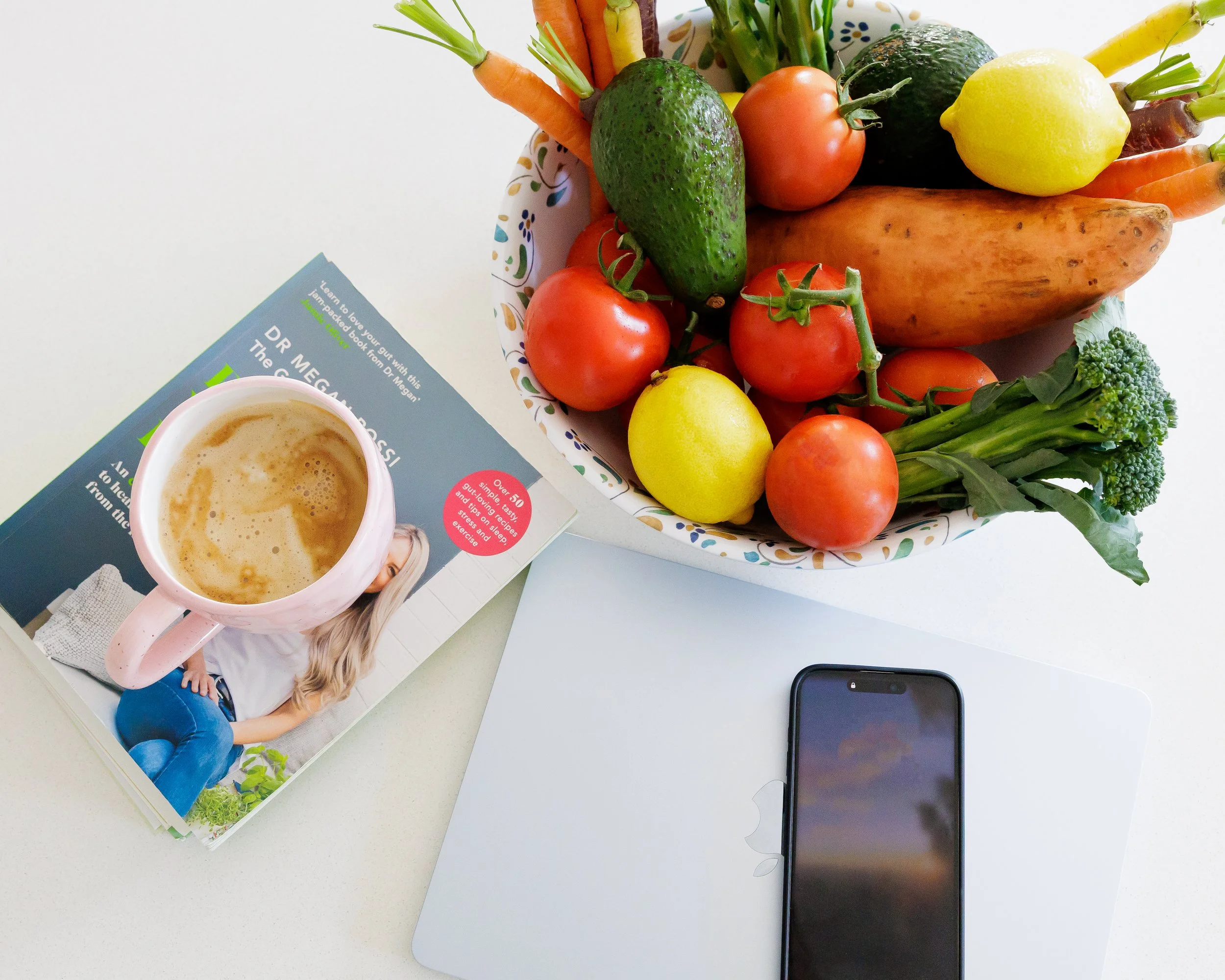A white table holding a bouquet of fresh vegetables including carrots, avocado, tomatoes, lemon, and broccoli. There is a magazine, a cup of coffee, and a smartphone also on the table.