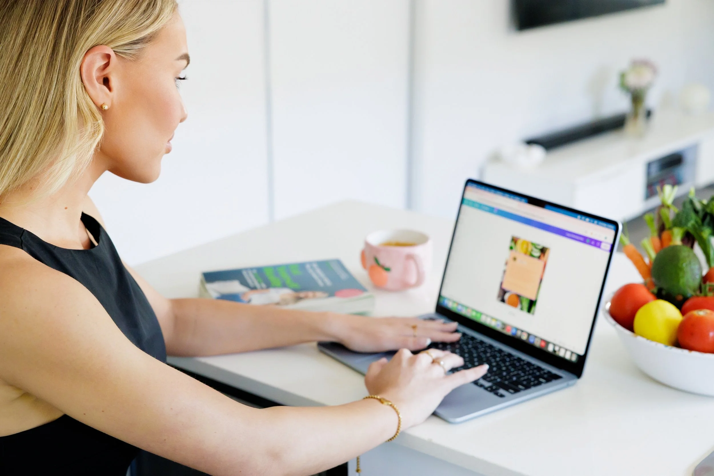 A woman with blonde hair working on a laptop at a white table, with a book, a pink mug, and a bowl of colorful vegetables nearby.