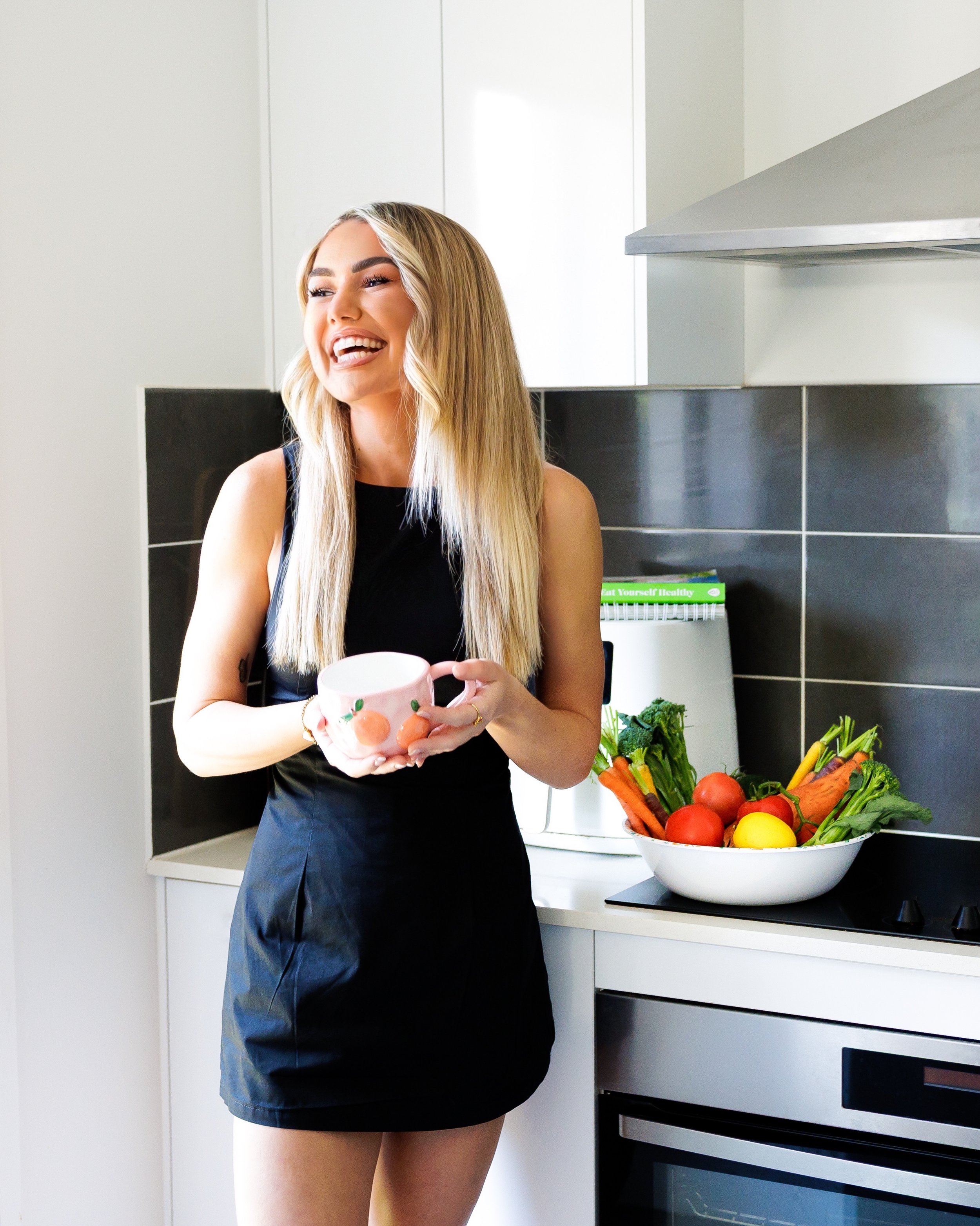 A young woman with long blonde hair smiling and holding a pink mug with a fruit design, standing in a modern kitchen with a wide bowl of fresh vegetables on the counter behind her.