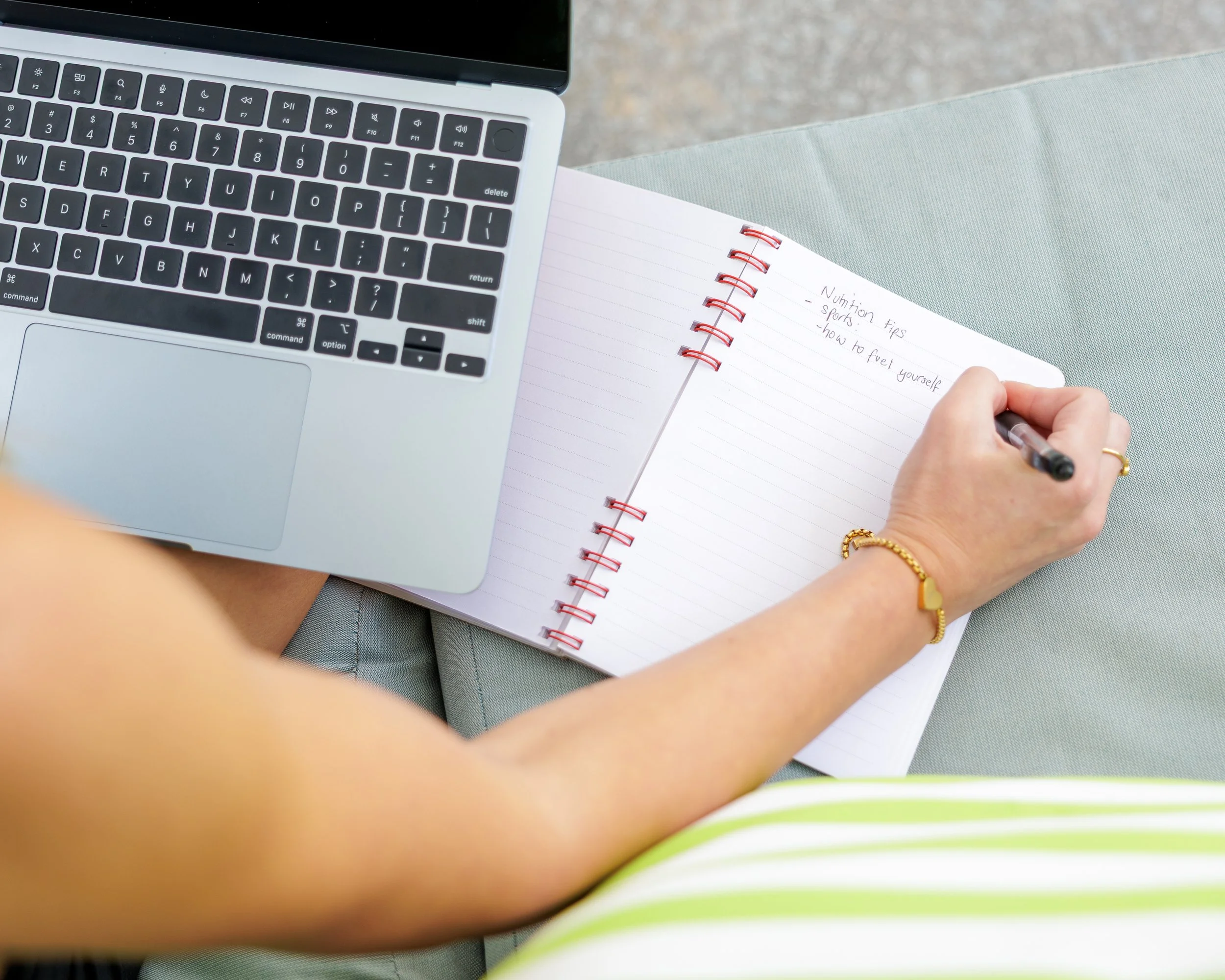 A person's hand holding a pen and writing in a spiral notebook with notes about nutrition tips, sports, and how to fuel yourself, next to a laptop on a surface.