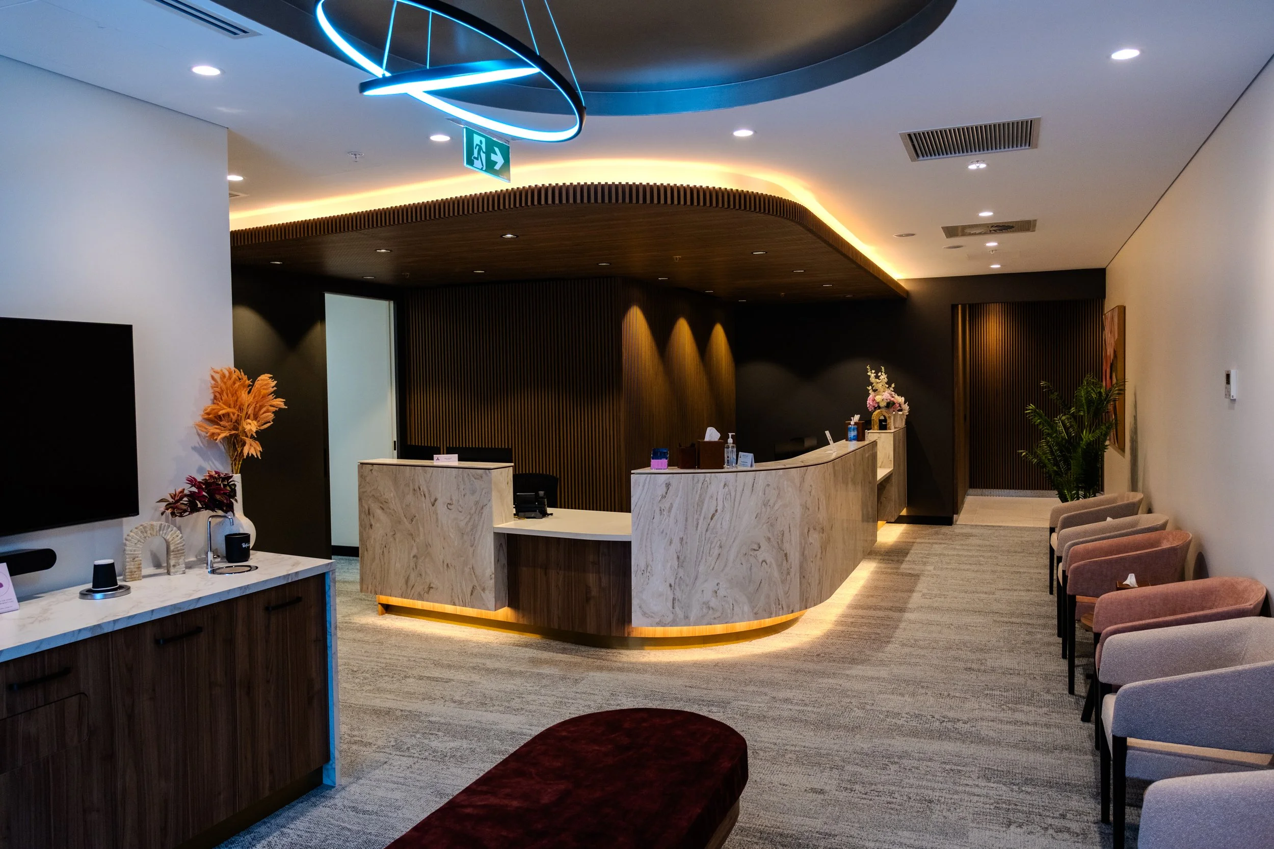Modern hotel lobby with a circular reception desk, beige marble, and wood accents. Stylish seating with pink and gray chairs, potted plants, flowers, and contemporary lighting fixtures.