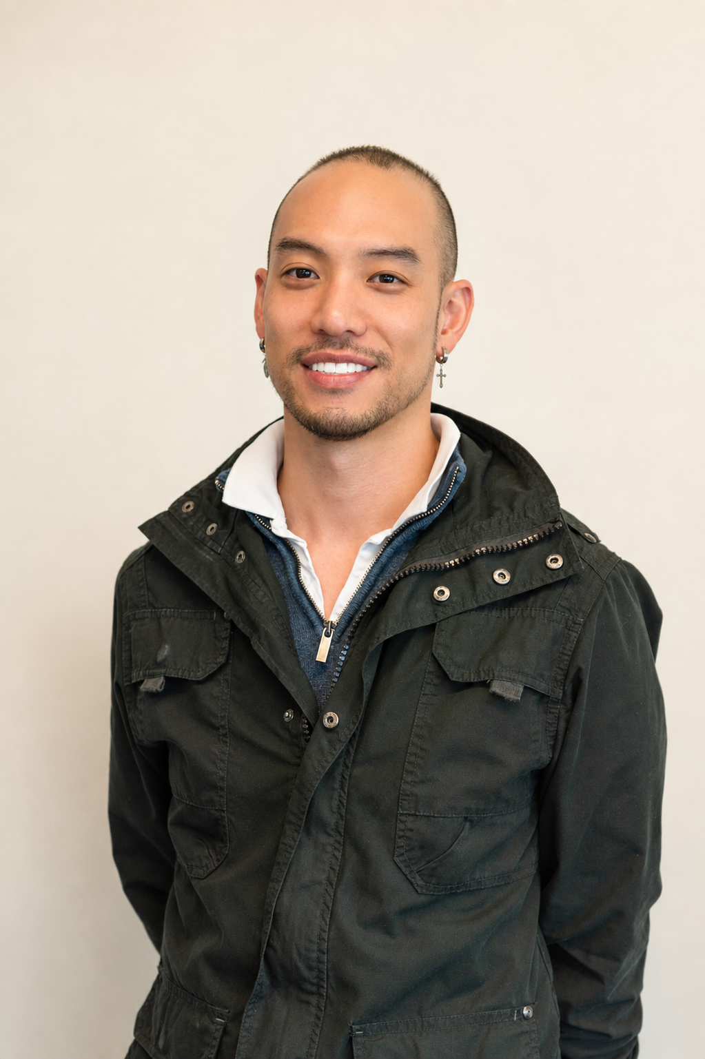 A young man with a shaved head, smiling, wearing a black jacket over a white collared shirt, standing against a plain white wall.