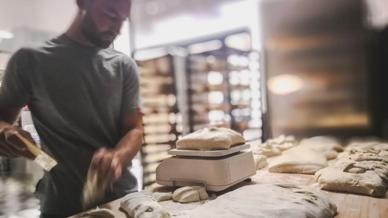 A person working with dough in a bakery kitchen with racks of baked goods in the background.