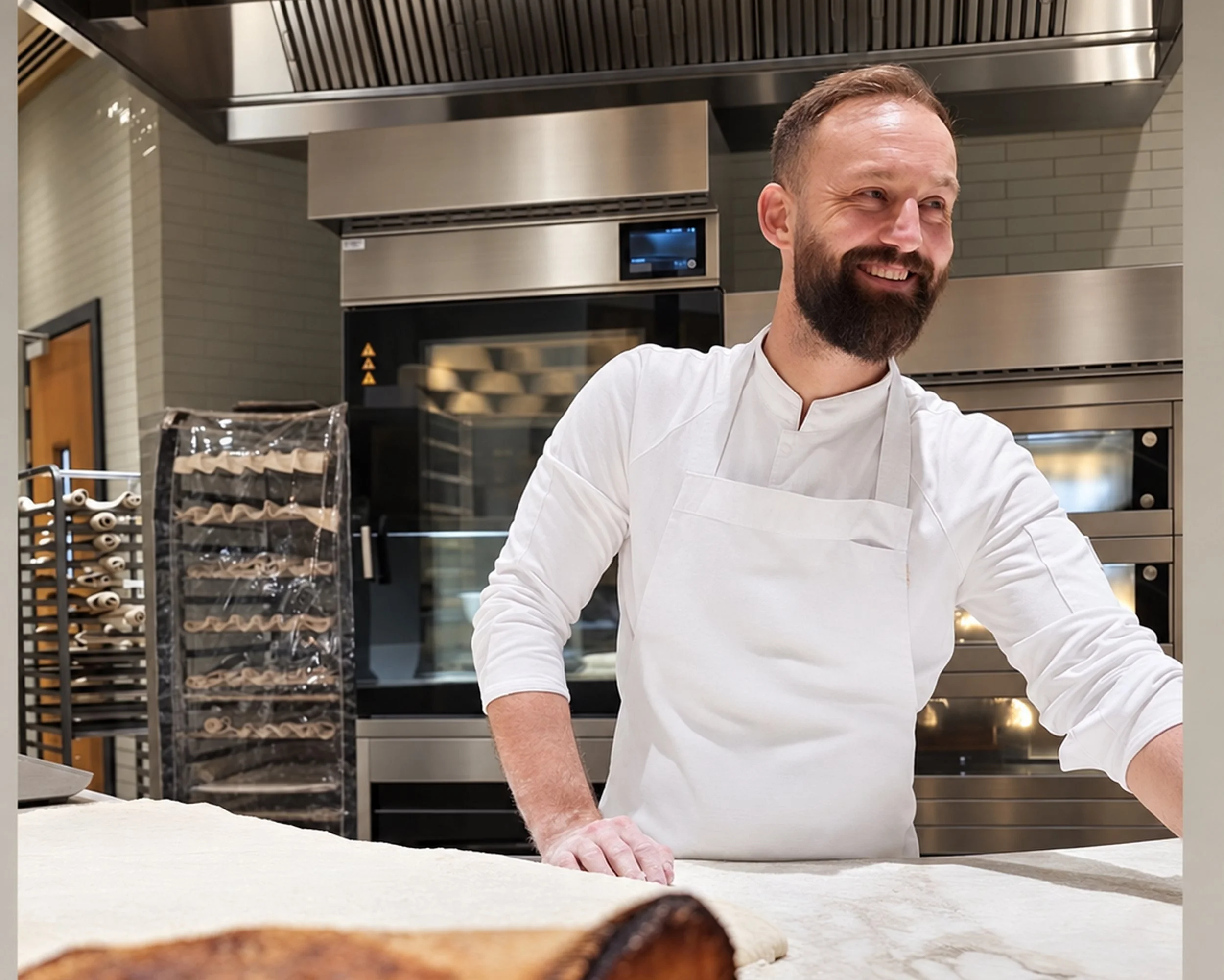 A male chef with a beard wearing a white chef's coat and apron smiles while working in a professional kitchen with stainless steel appliances and racks of baked goods.