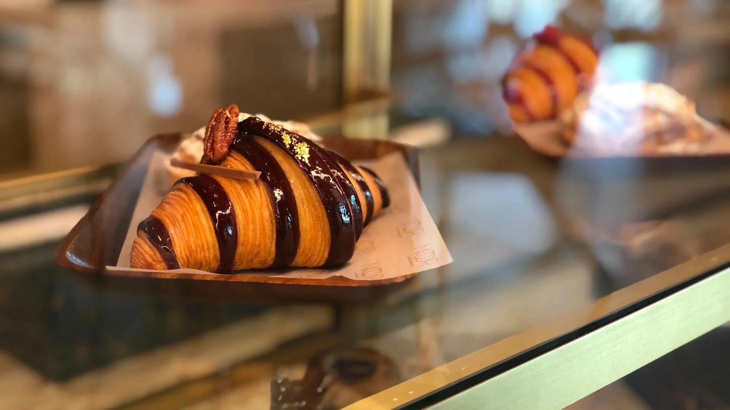 A croissant topped with chocolate drizzle and a pecan, placed on a paper-lined tray in a display case.