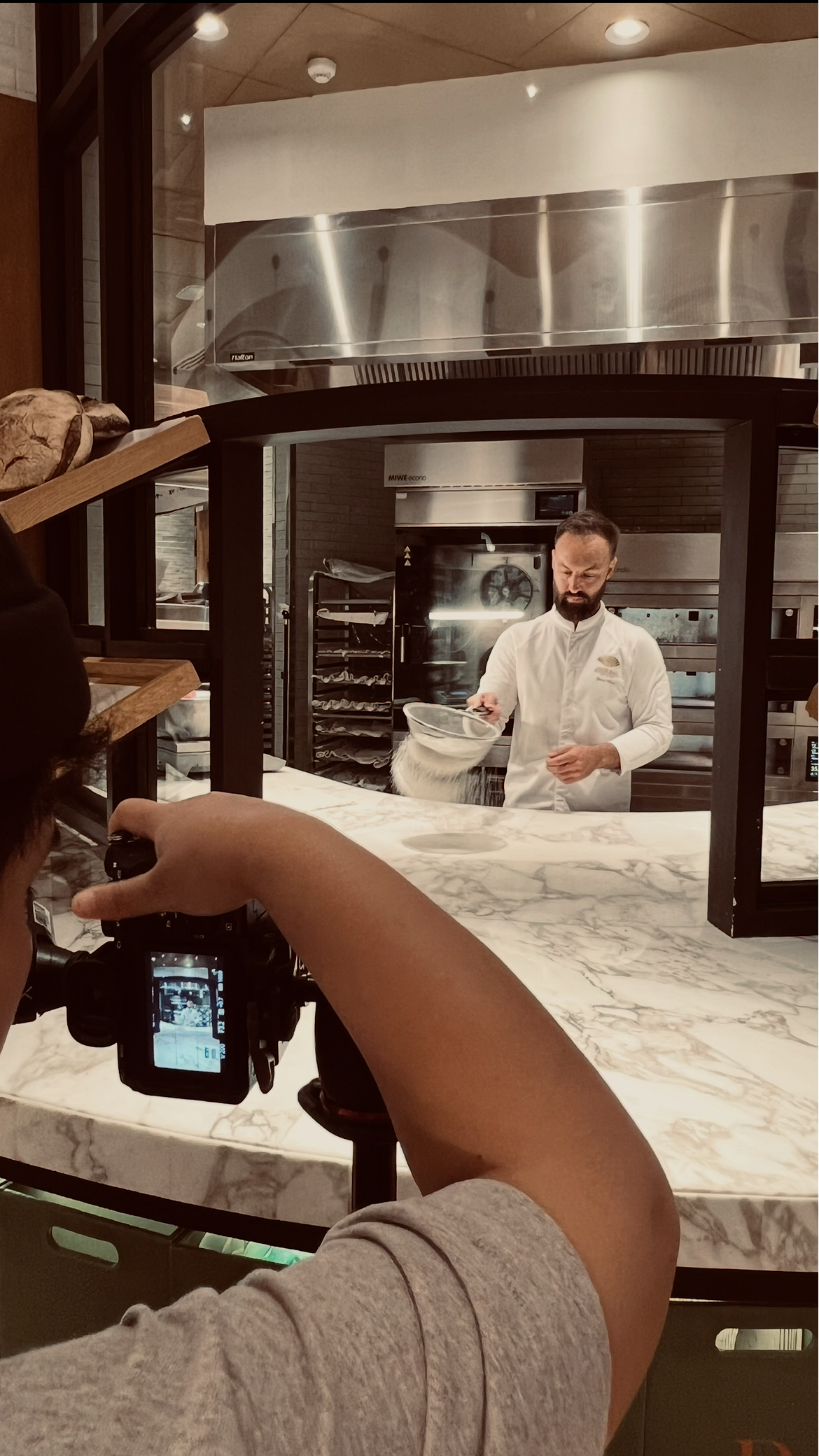 A chef wearing a white uniform is flipping a pizza dough in the air inside a modern restaurant kitchen. The scene is viewed through a glass window, with a person in a gray shirt holding a camera in the foreground. The kitchen has stainless steel appliances and a marble counter.