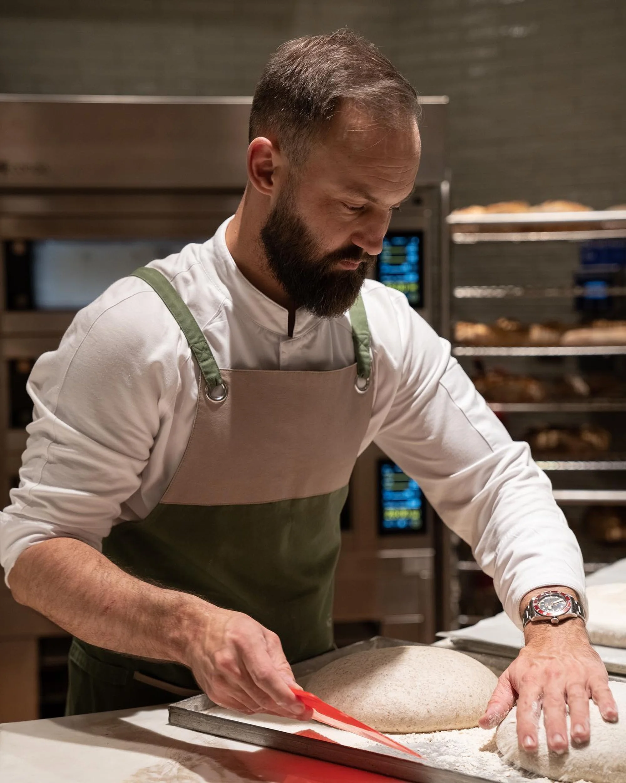A man with a beard and a wristwatch is kneading dough on a baking tray, wearing a white shirt and green apron, in a professional kitchen with racks of bread in the background.