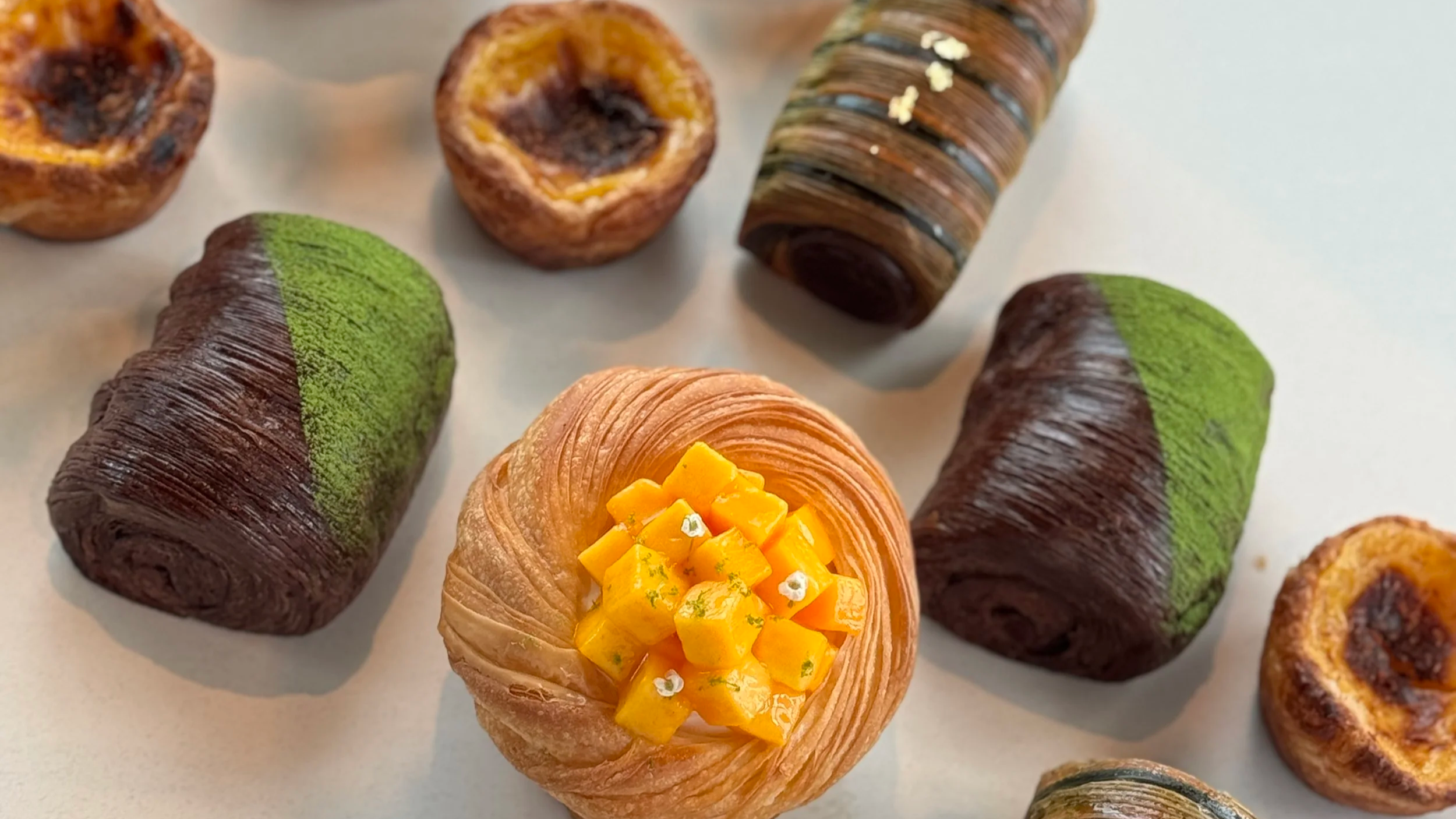 A variety of colorful pastries, including rolled layered pastries with green and brown icing, a pastry topped with small yellow fruit cubes and white edible flowers, and other pastries with different fillings and textures on a plain background.