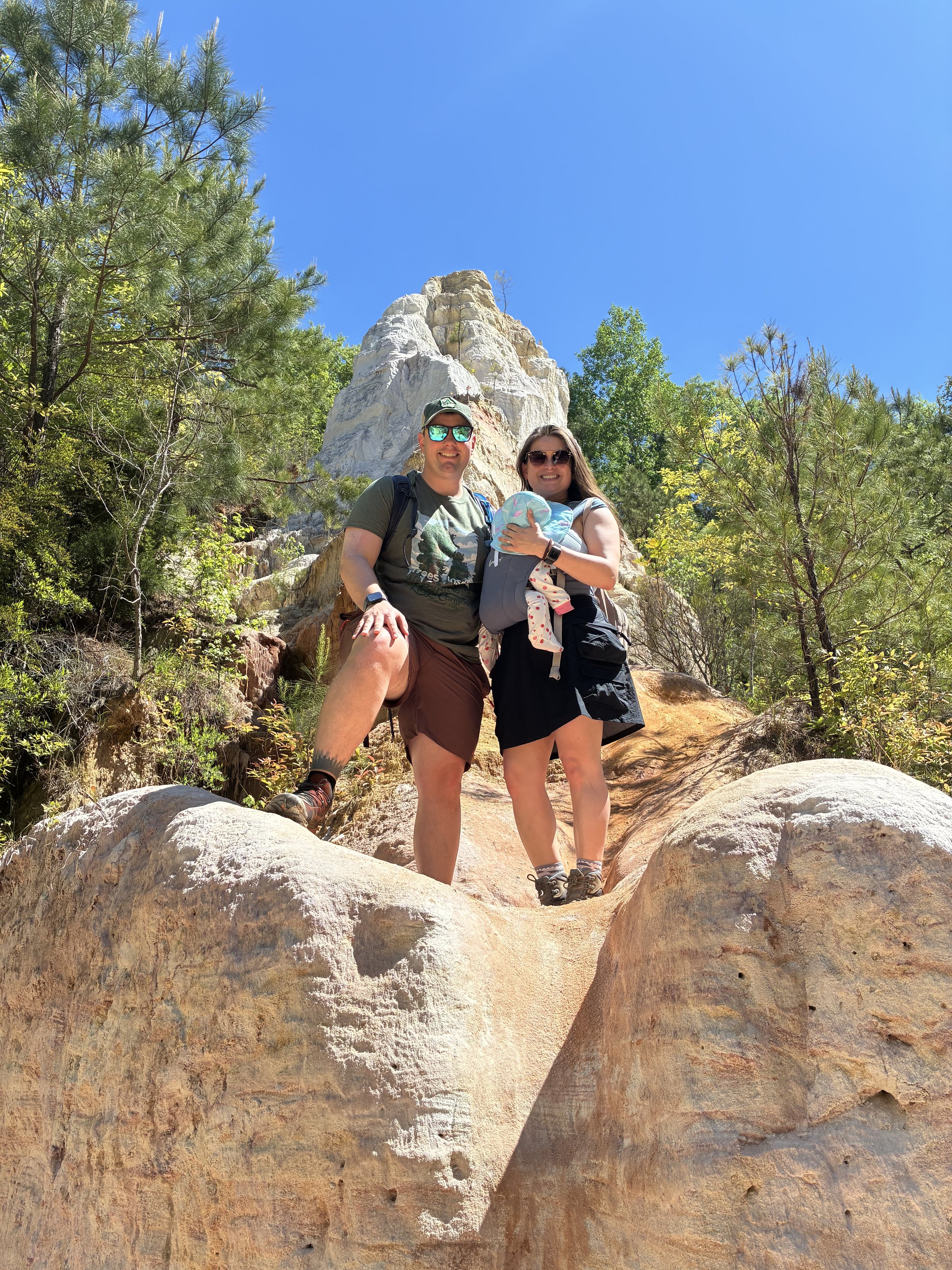 A man and woman hiking on a rocky trail outdoors, with a large white rock formation and green trees behind them, under a clear blue sky.