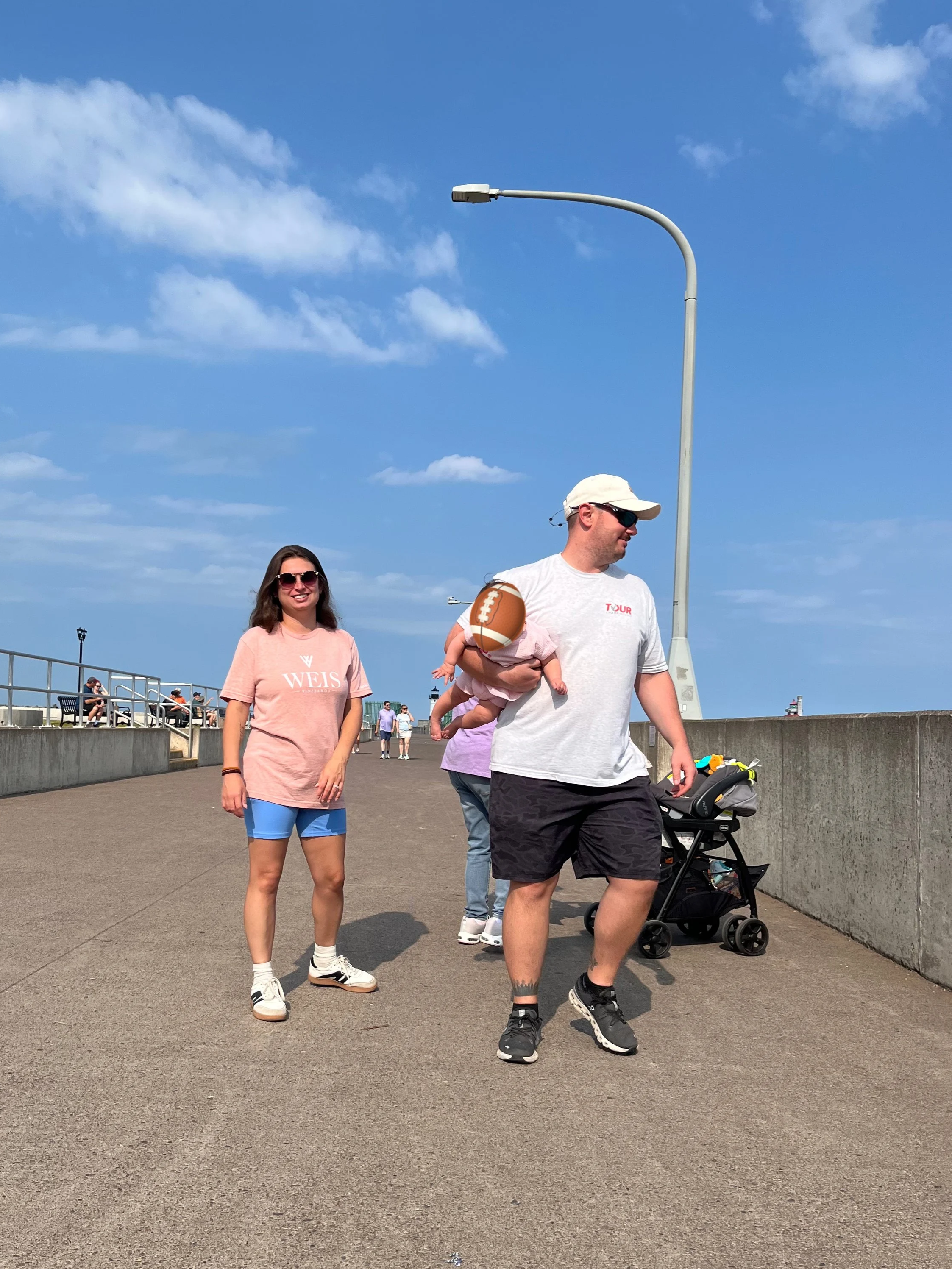 A man holding a baby with a football in a stroller walking on a concrete path, a woman wearing sunglasses and a pink shirt walking beside him, with a blue sky, a street lamp, and other people sitting on benches in the background.