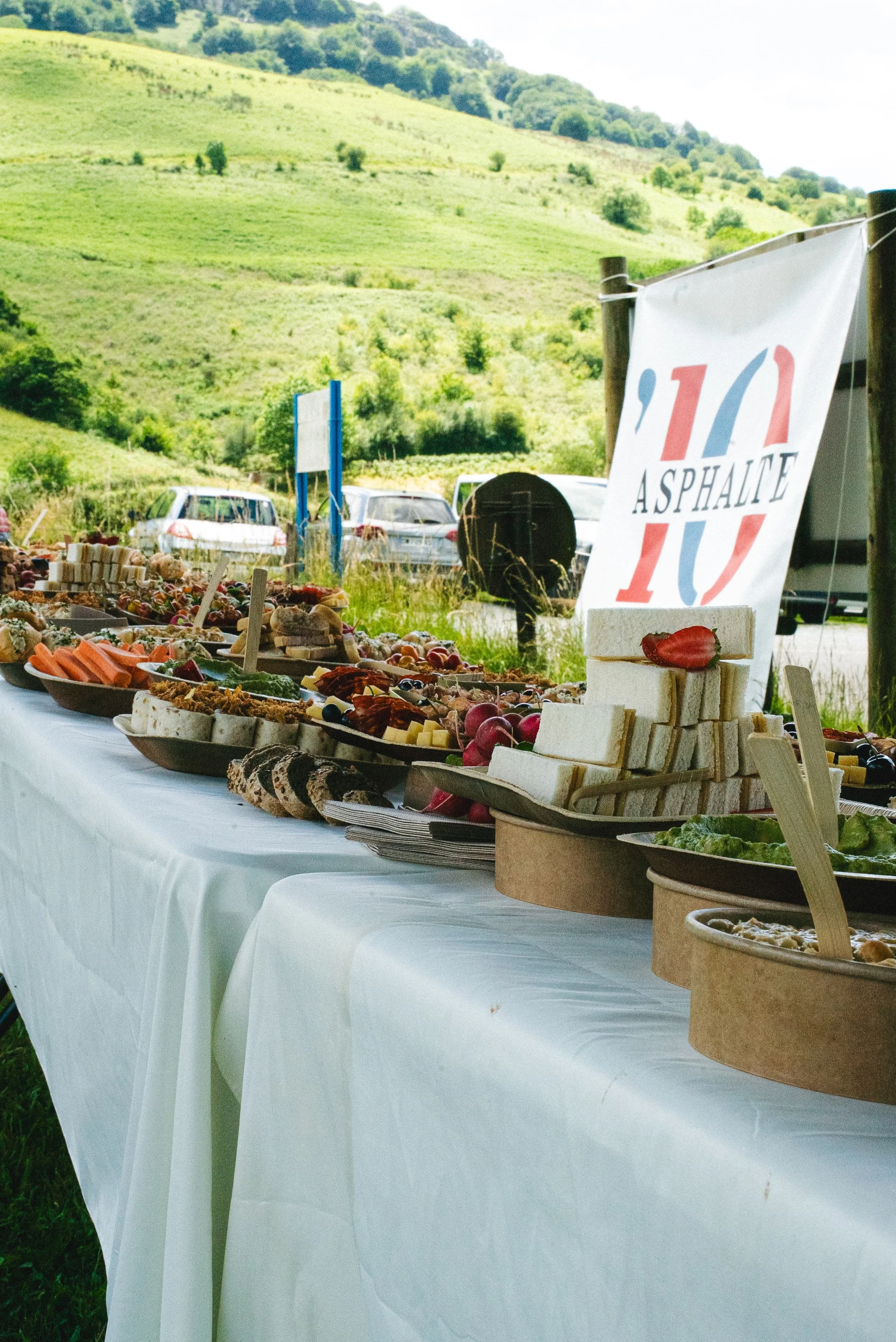 Table de plats et de fruits de saison à un marché en plein air avec collines verdoyantes en arrière-plan, affichant 'l'Asphalte 10'