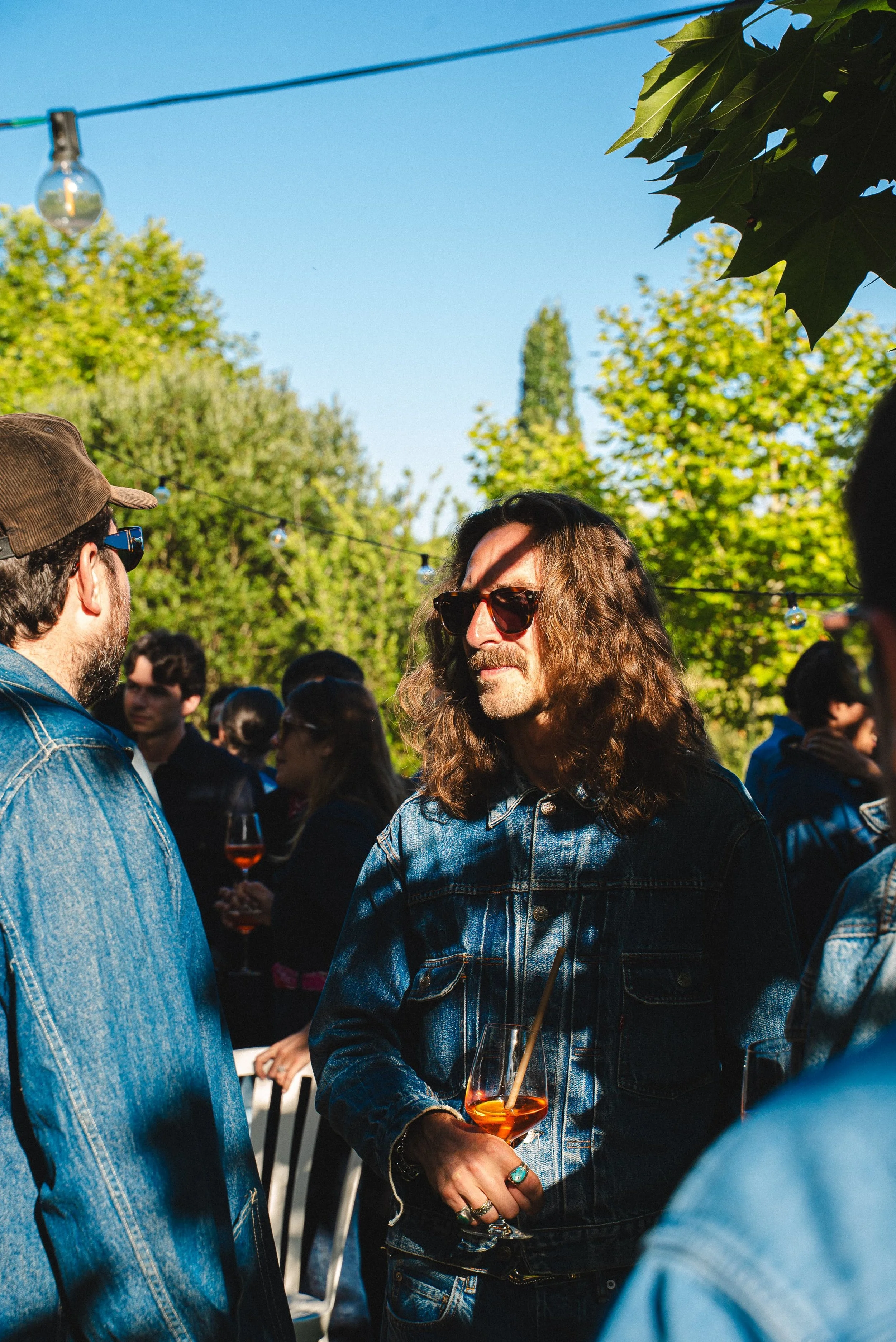 Une personne avec des cheveux longs et ondulés, portant des lunettes de soleil et un jean en interaction lors d'un événement extérieur avec un autre individu portant une casquette et des lunettes.