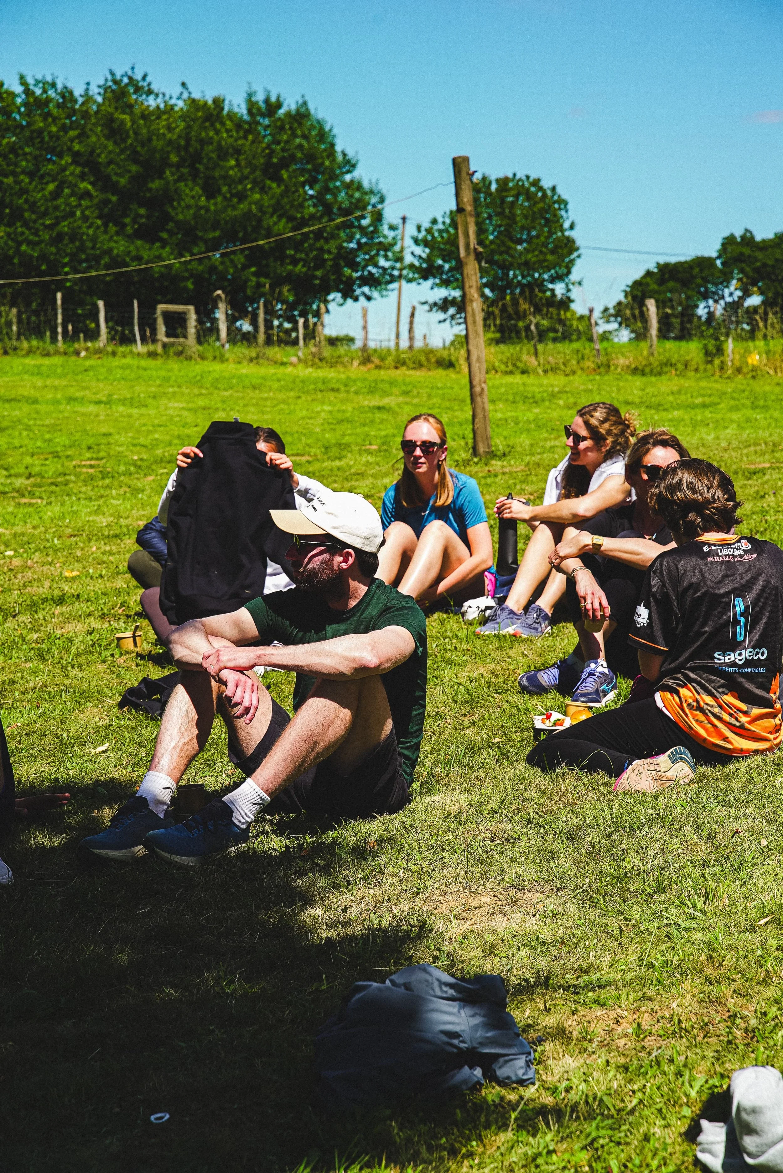 Groupe de personnes assises sur l'herbe dans un champ en plein air, avec des arbres et un ciel bleu en arrière-plan, probablement lors d'une activité de plein air ou d'une sortie en groupe.