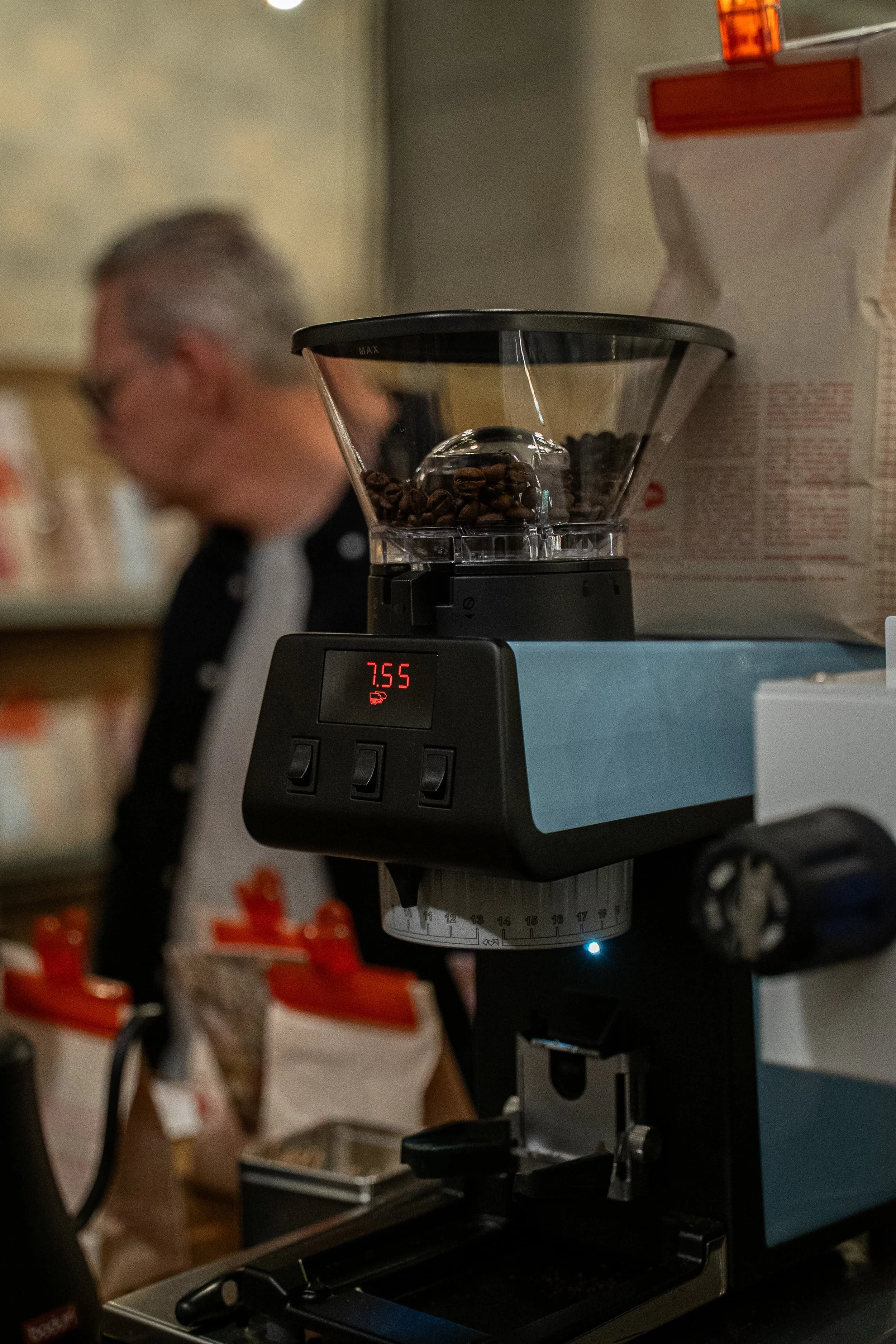 Un moulin à café en train de moudre des grains de café. En arrière-plan, on voit une personne floue portant des lunettes et un tablier noir.