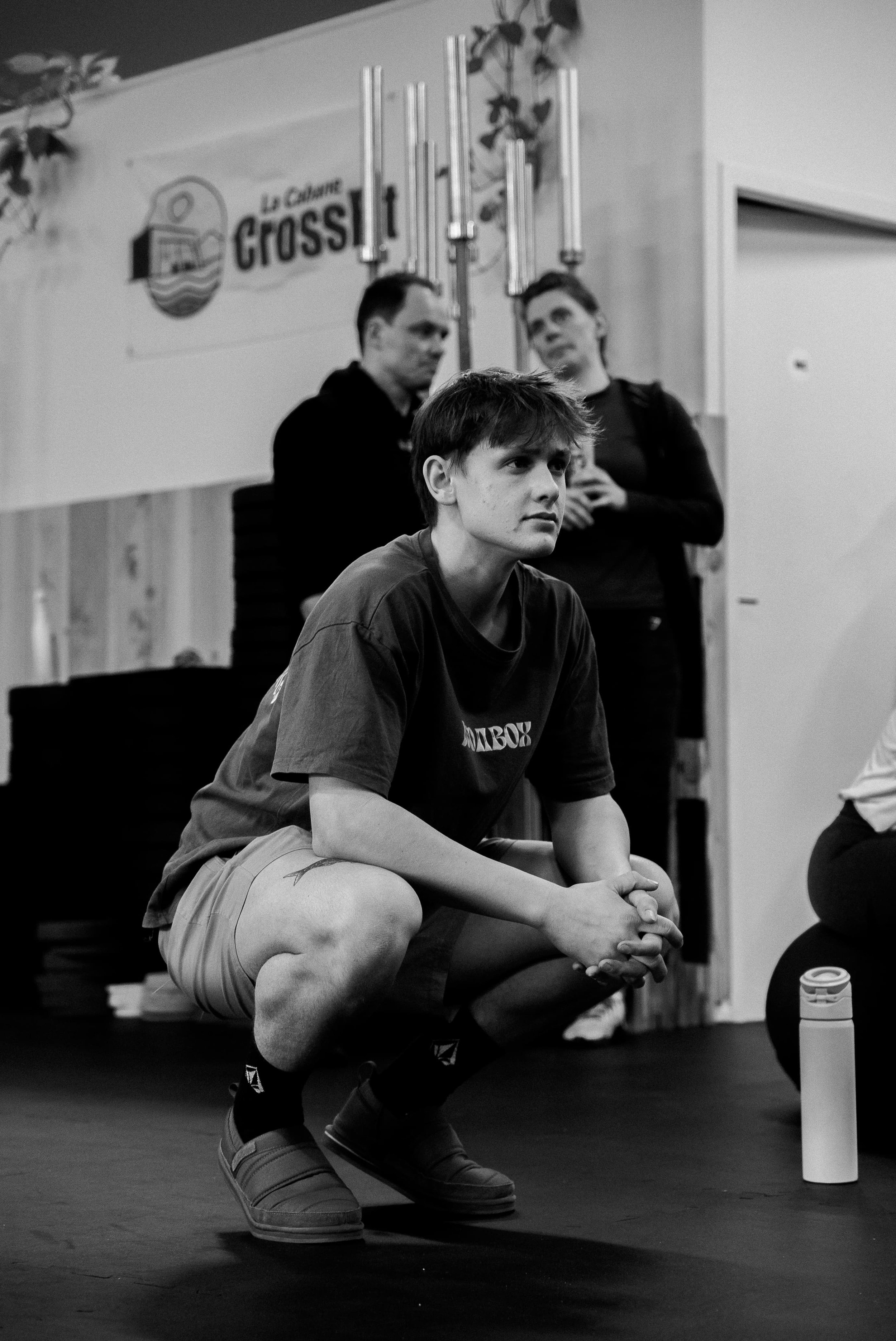Un jeune homme assis en position accroupie dans une salle d'entraînement, portant un t-shirt et des shorts, avec deux autres personnes en arrière-plan près d'un mur avec un logo, et une bouteille d'eau à proximité.