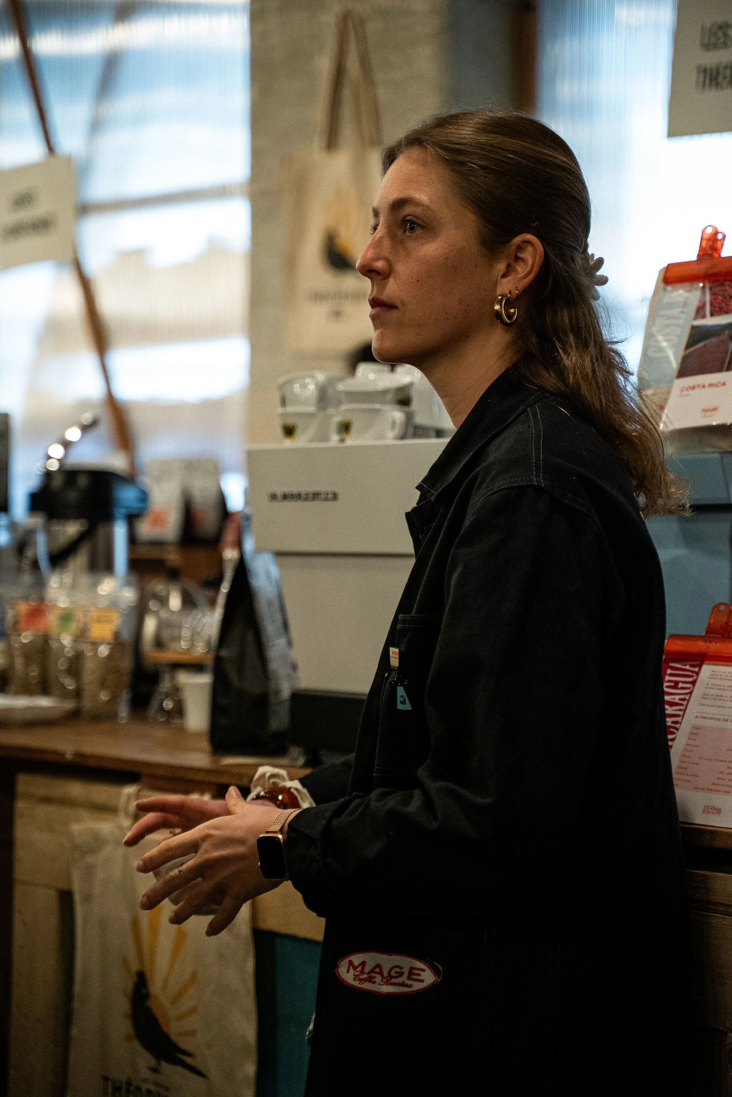 Une femme avec des cheveux bruns attachés, portant des boucles d'oreilles en or et une montre connectée, qui se tient à l'intérieur devant un comptoir ou une caisse, peut-être dans une boutique ou un café.