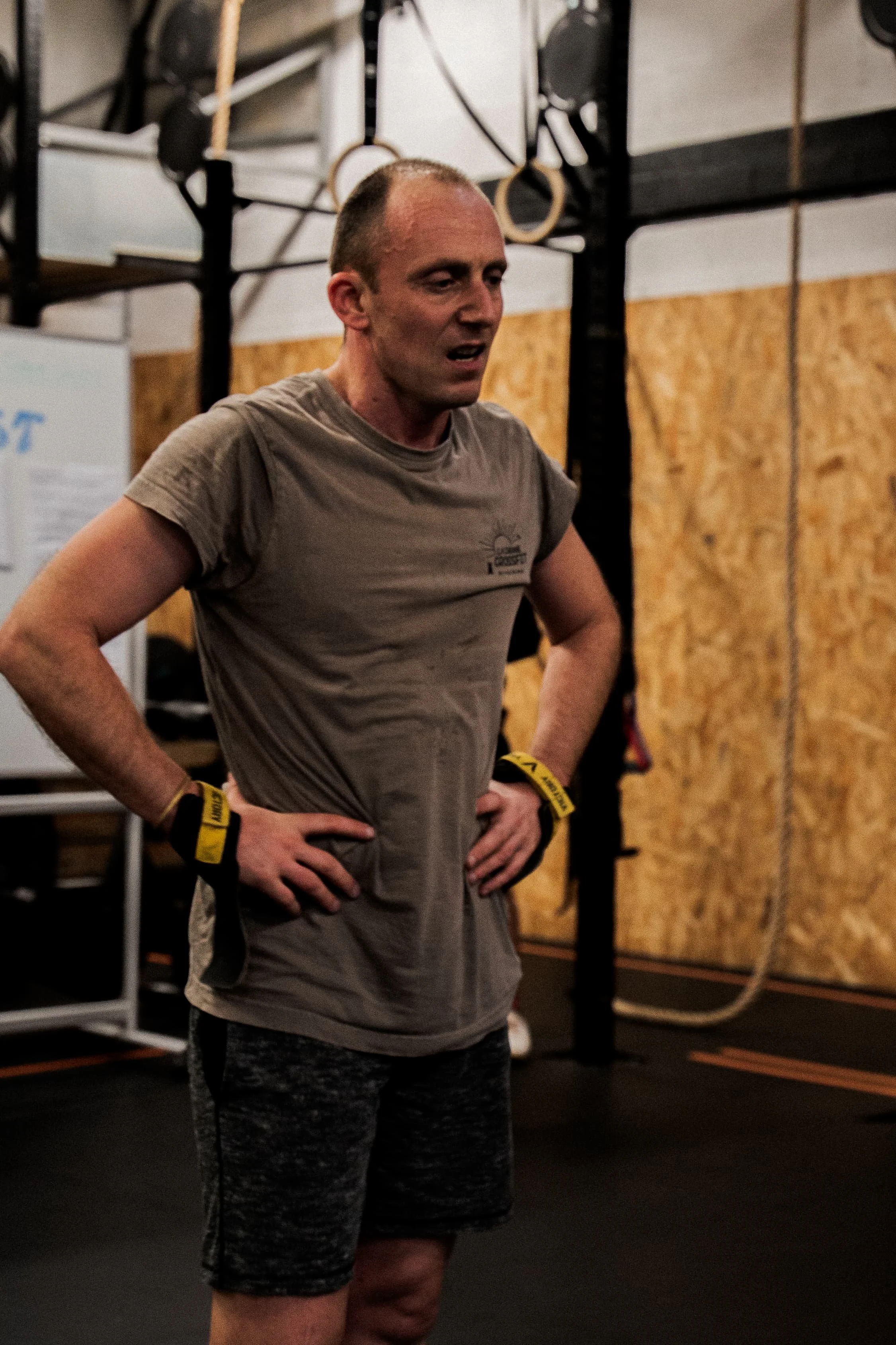 Un homme dans une salle de sport, portant un t-shirt gris et des vêtements de sport, avec des poids et des équipements d’entraînement en arrière-plan.