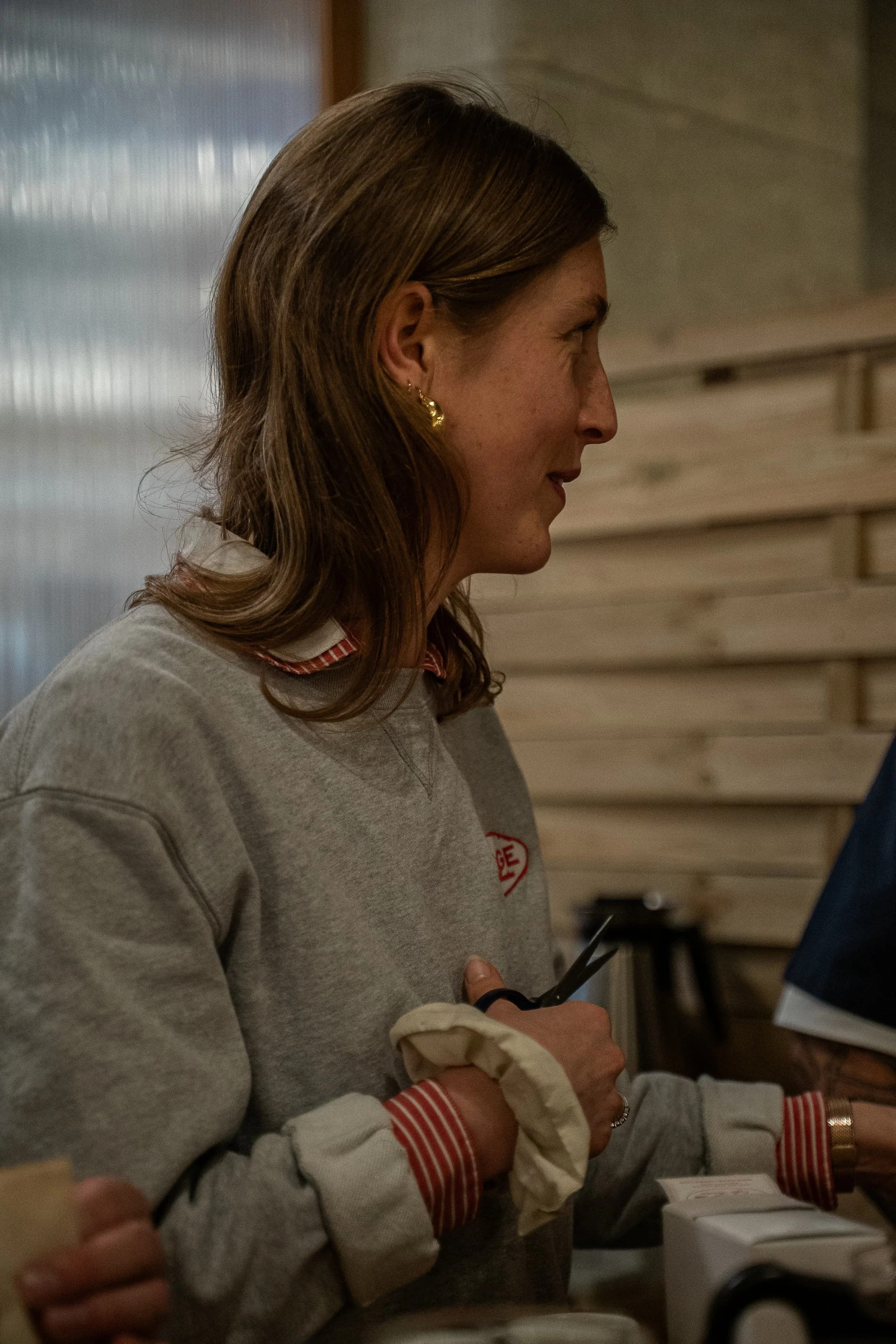 Une femme avec des cheveux bruns longs, portant un sweat gris avec un col à rayures rouges et blanches, tenant un chiffon blanc dans ses mains.