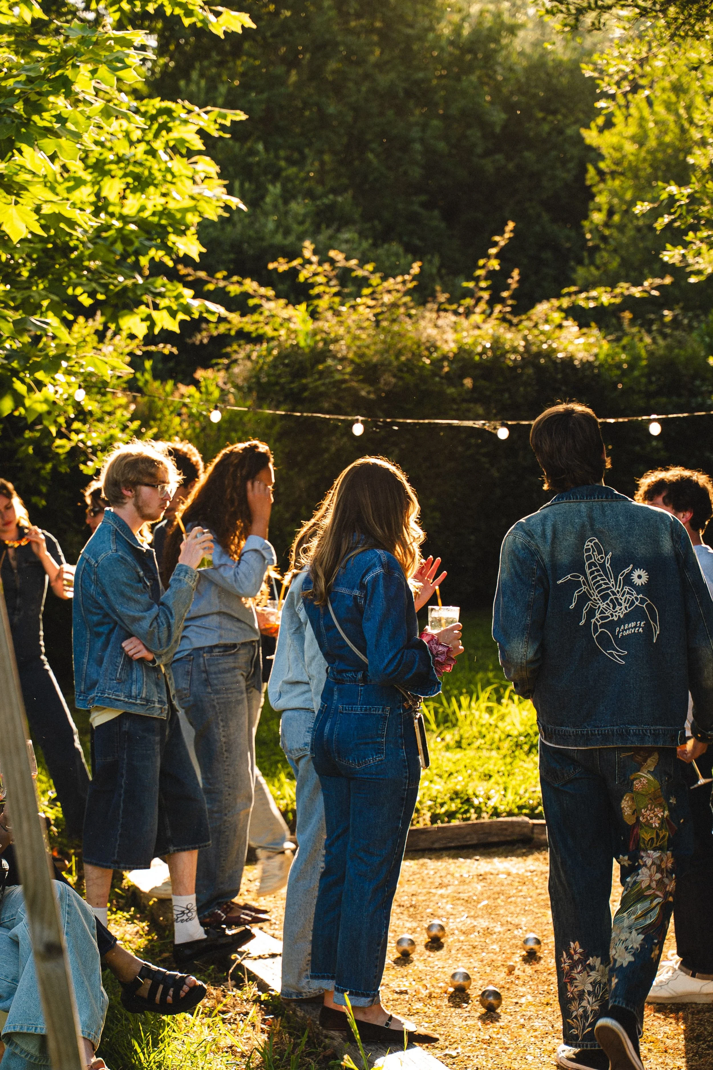 Groupe de personnes jouant à la pétanque dans un jardin en plein air, avec des arbres verts en arrière-plan et des guirlandes lumineuses suspendues au-dessus.
