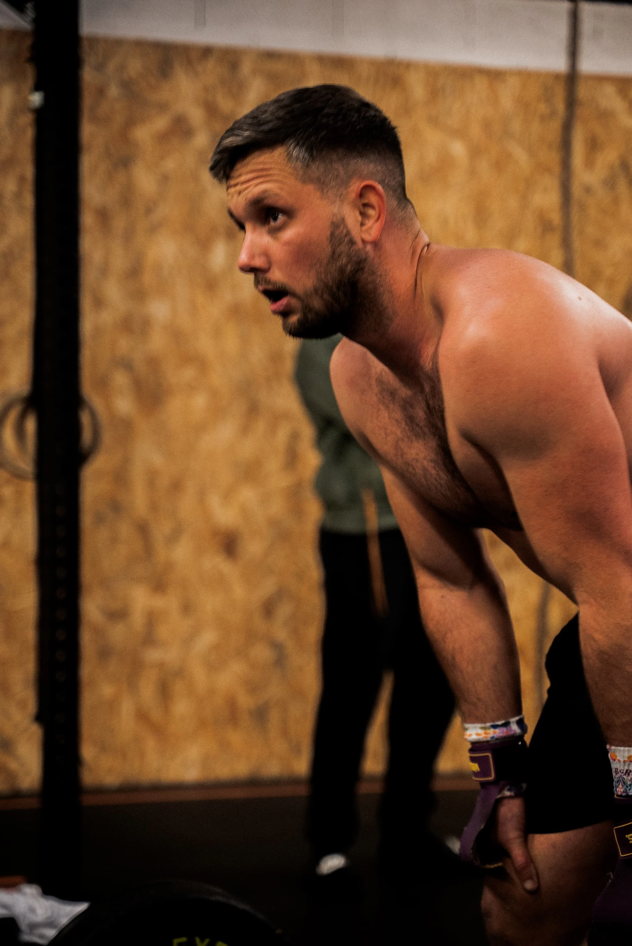 Homme musclé, sans vêtement supérieur, en train de s'entraîner avec des poids ou dans une salle de sport, avec un mur en bois en arrière-plan.