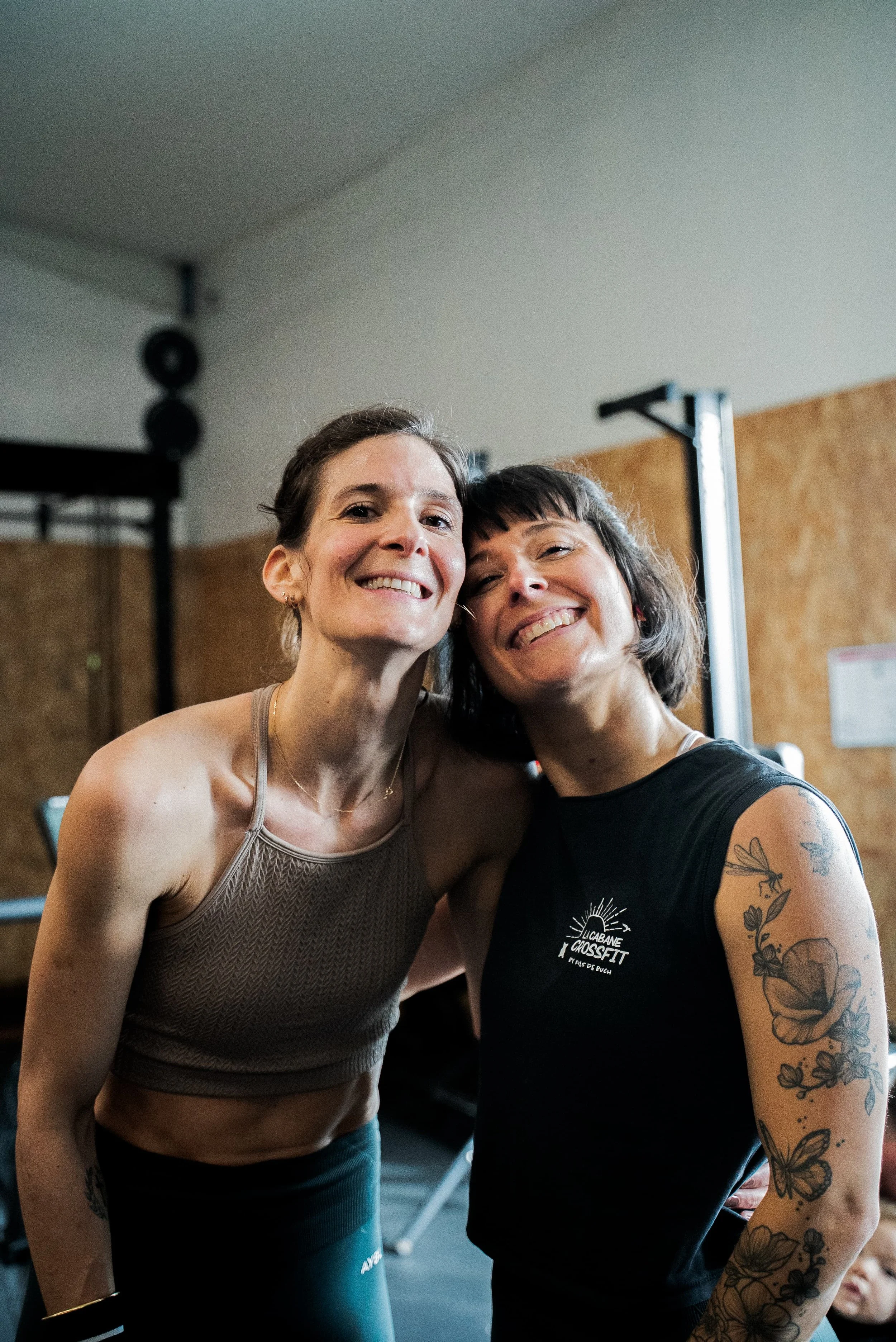 Deux femmes souriantes posent ensemble dans une salle de sport, l'une porte un débardeur beige et l'autre un t-shirt noir avec un logo de CrossFit, affichant des tatouages sur le bras.