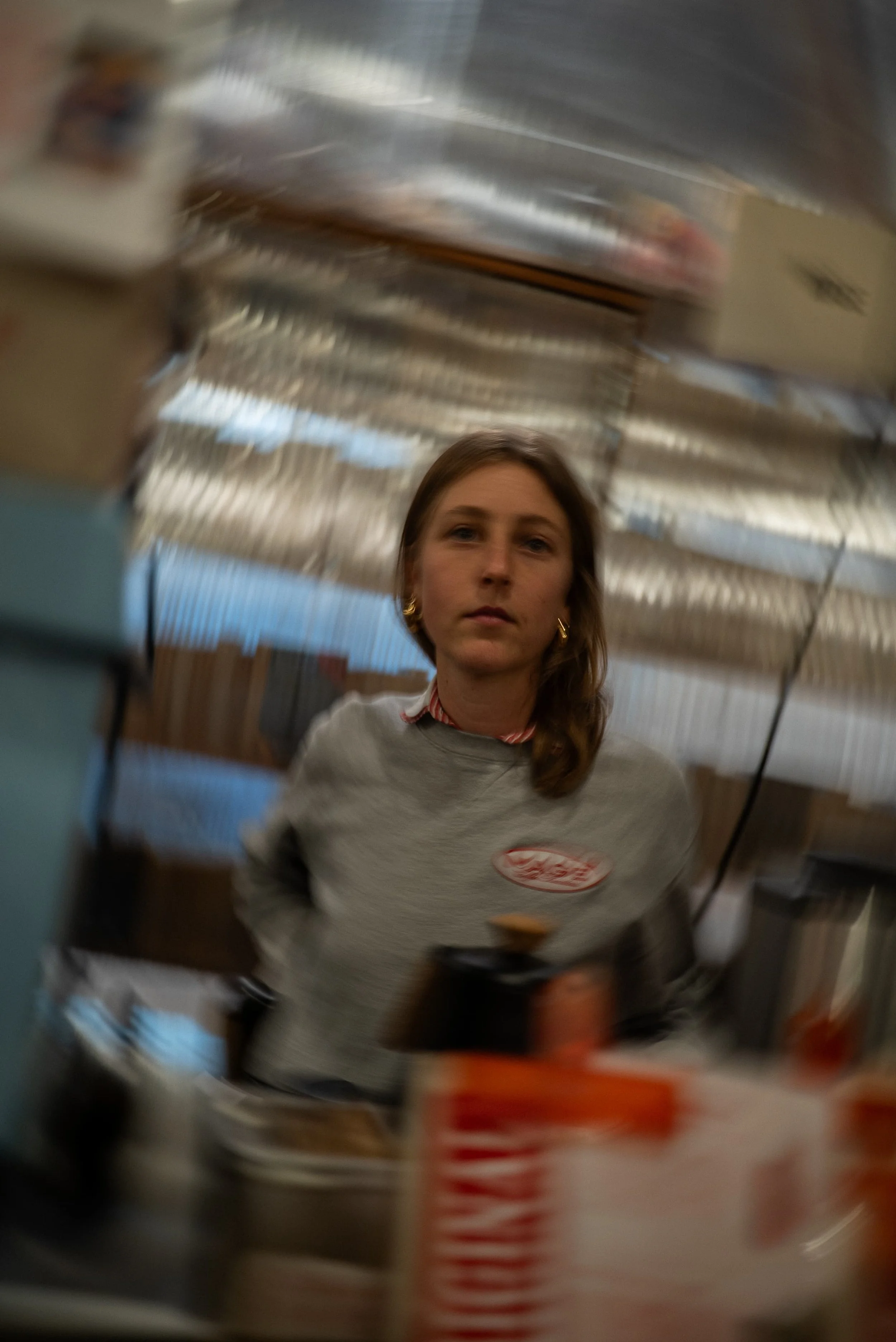Une femme avec des cheveux bruns et une chemise à col rouge et blanc qui regarde vers la caméra, prise à travers une surface courbée ou un objet rond, ce qui crée une perspective déformée.