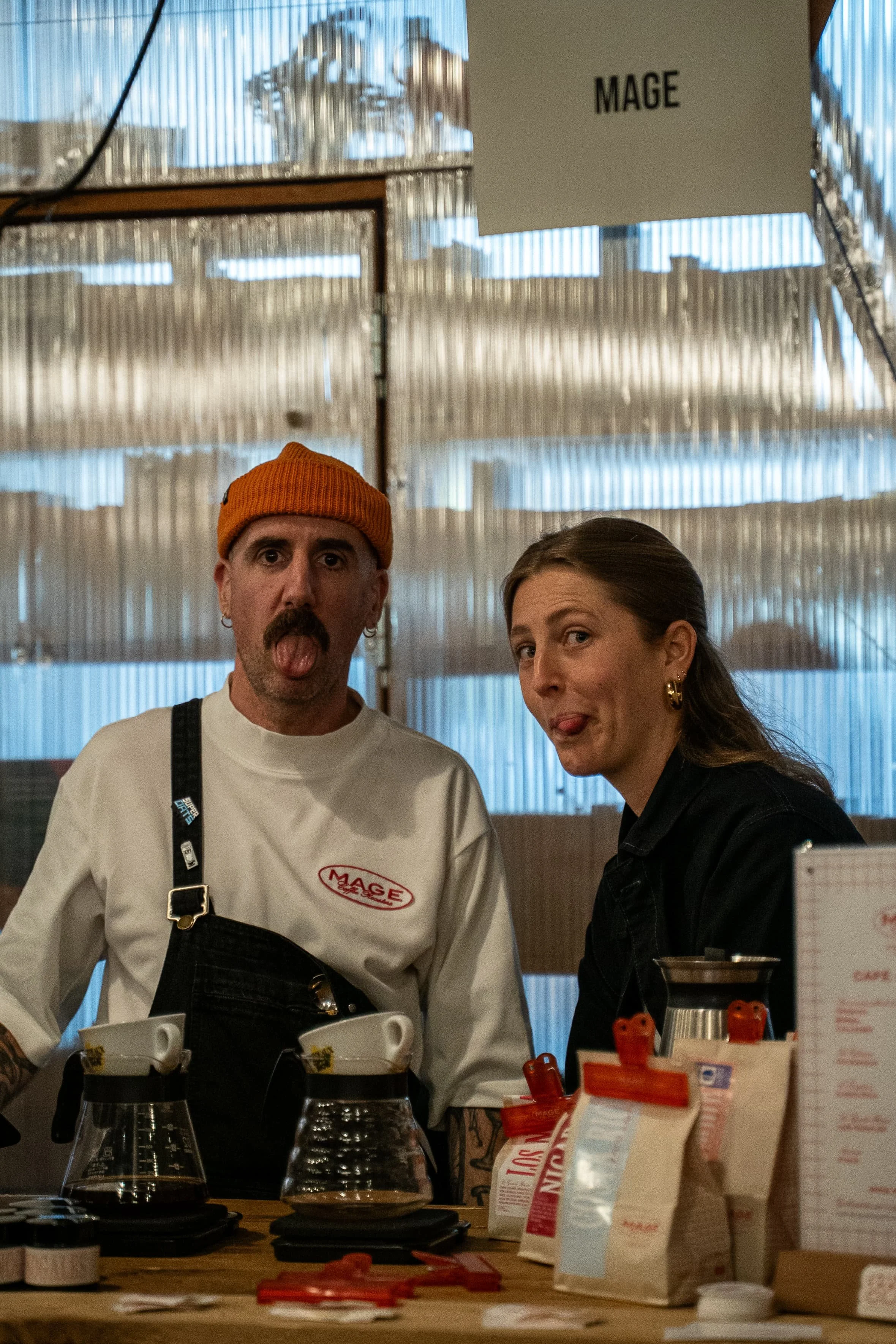Deux personnes, un homme avec un bonnet orange et une femme avec des boucles d'oreilles dorées, en train de prendre un café dans un café.