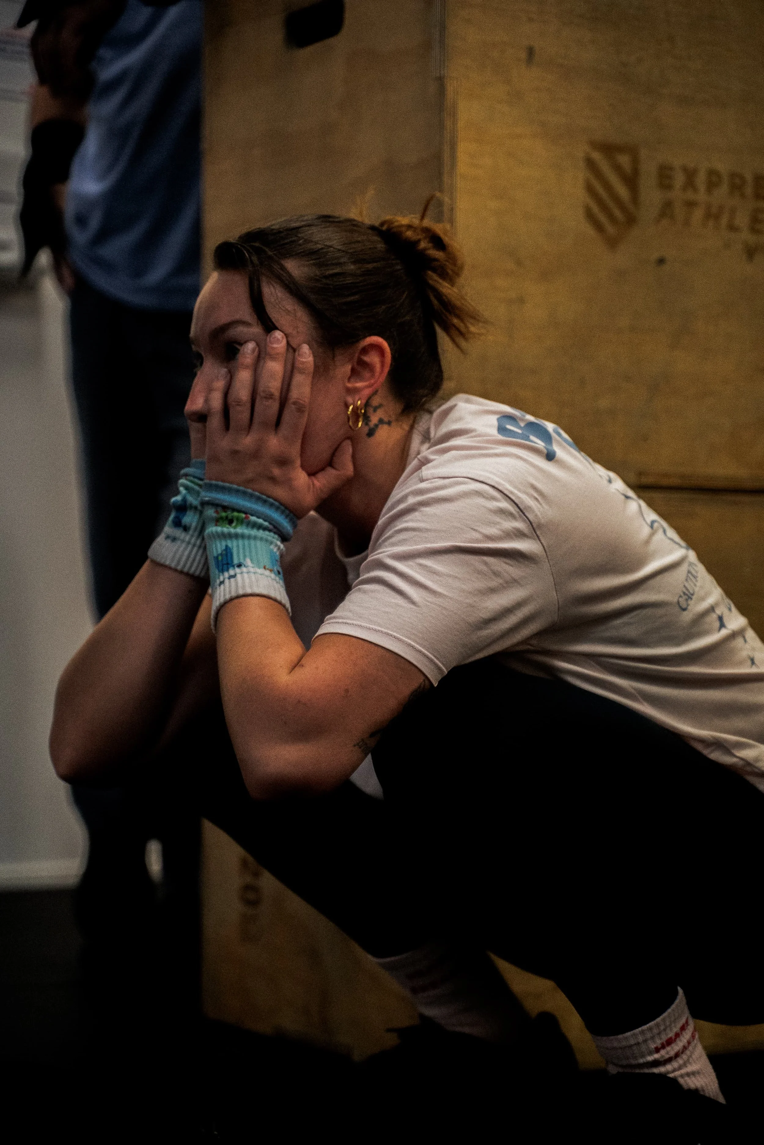 Une femme avec des cheveux attachés, portant un t-shirt blanc avec des motifs bleus, des bracelets de différentes couleurs, et des bijoux en or, est assise avec la main sur le visage dans une pièce avec un mur en bois et une boîte en carton à côté.