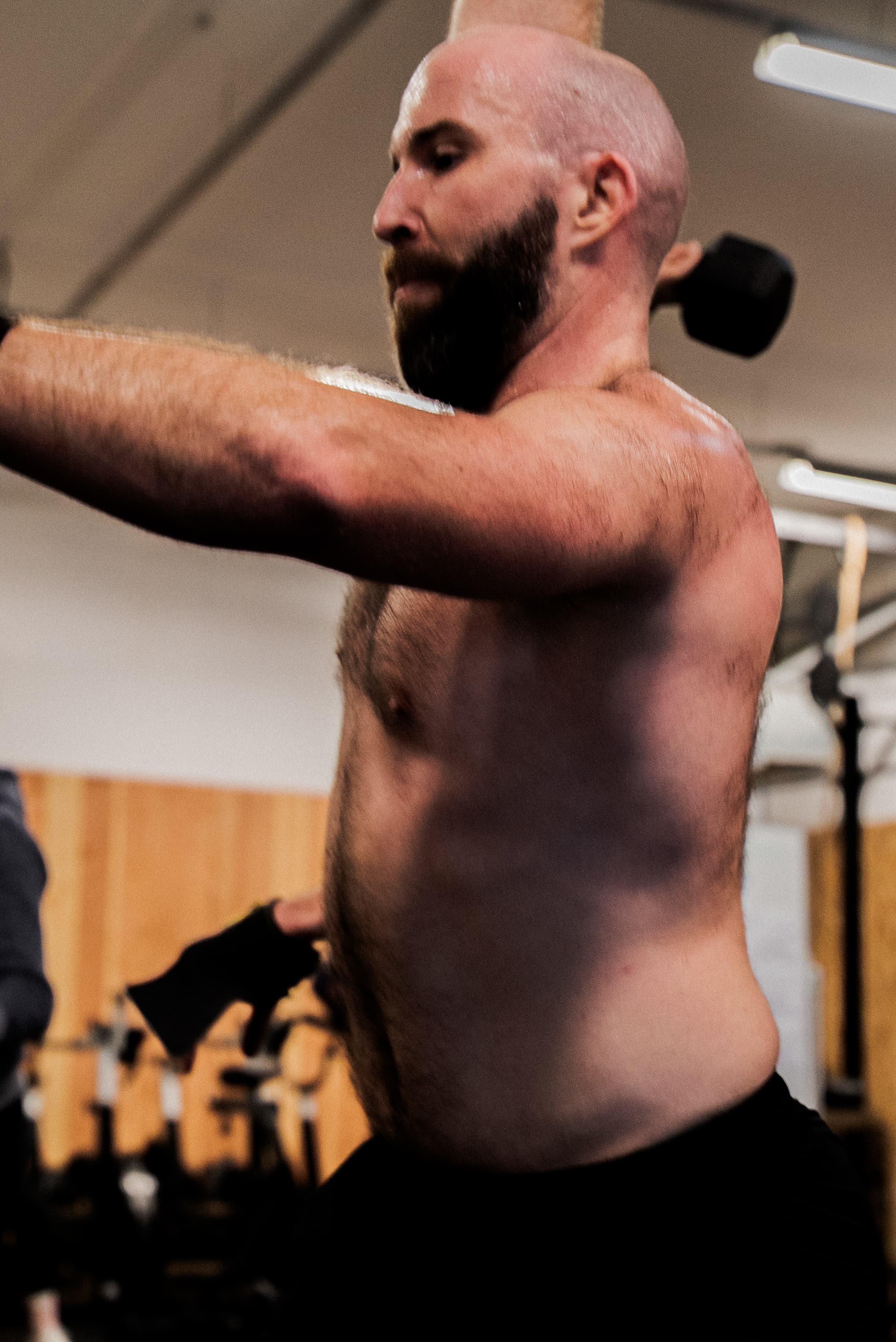 Homme barbu et chauve levant ses bras dans une salle de sport, portant un sweat noir, avec équipements de musculation en fond.