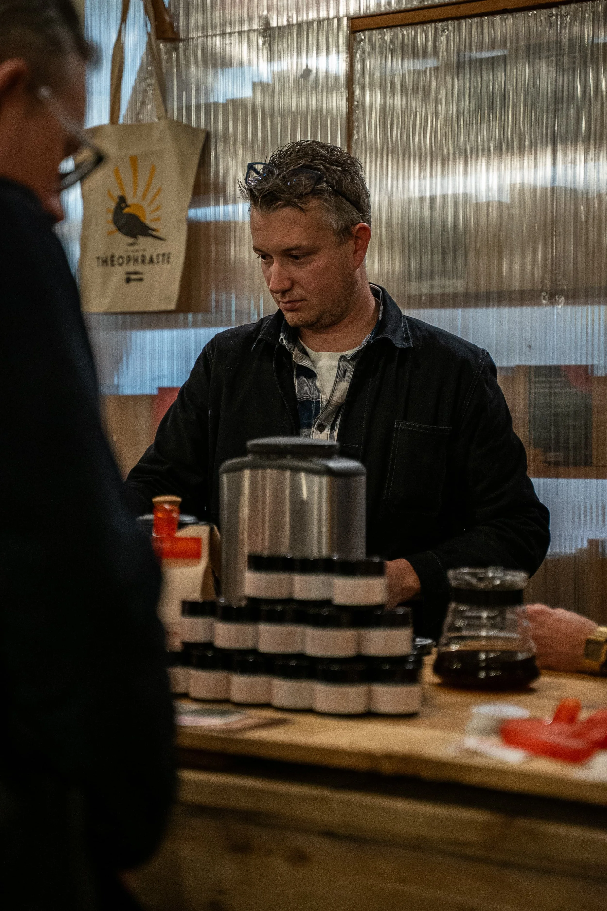 Jeune homme portant des lunettes sur la tête, assis derrière un comptoir dans un café ou une boutique, avec des piles de tasses et des objets en arrière-plan.