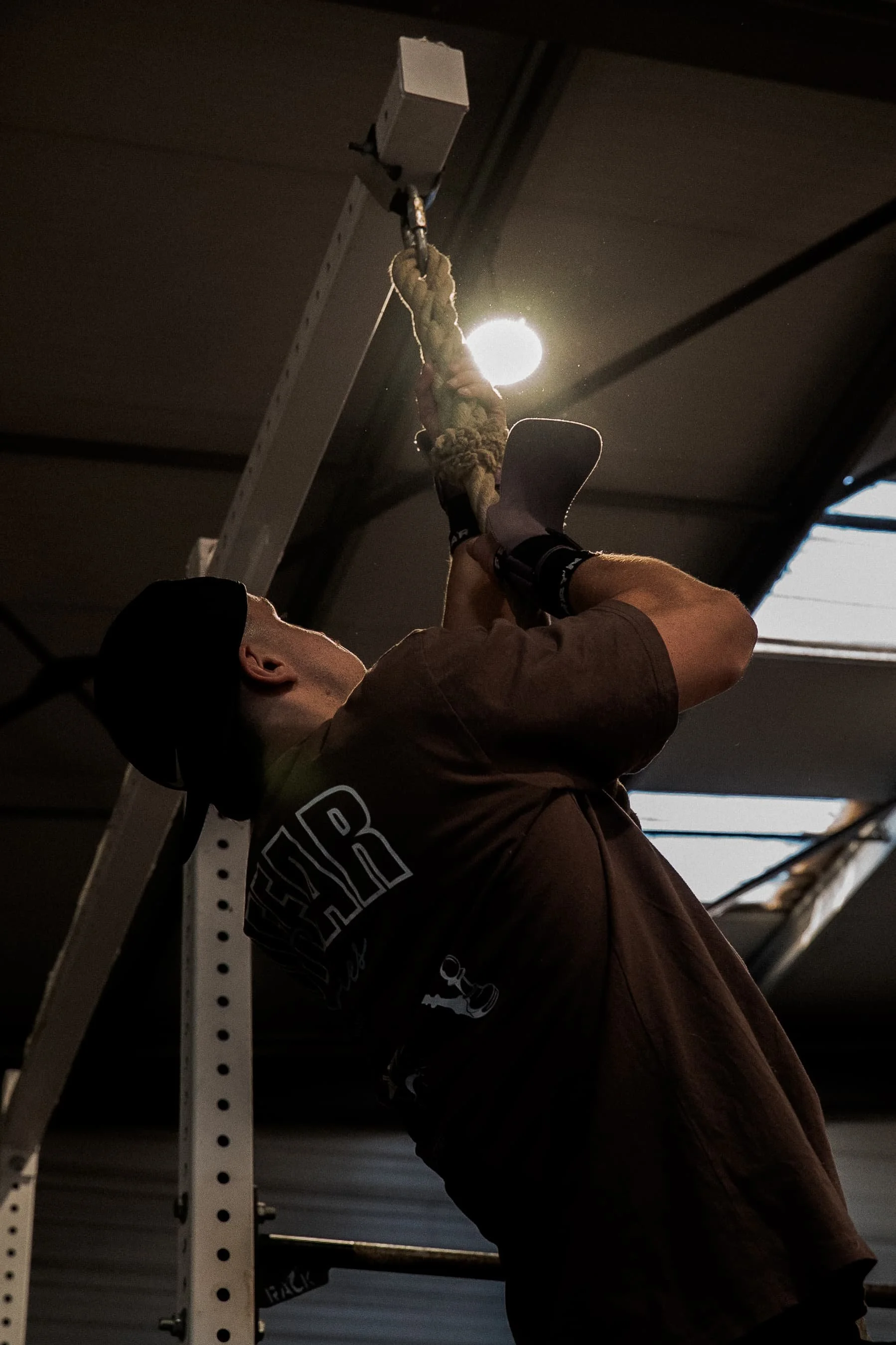 Une personne en train de faire de la musculation, suspendue à une barre de traction dans un garage ou une salle de sport, avec la lumière du soleil en arrière-plan.