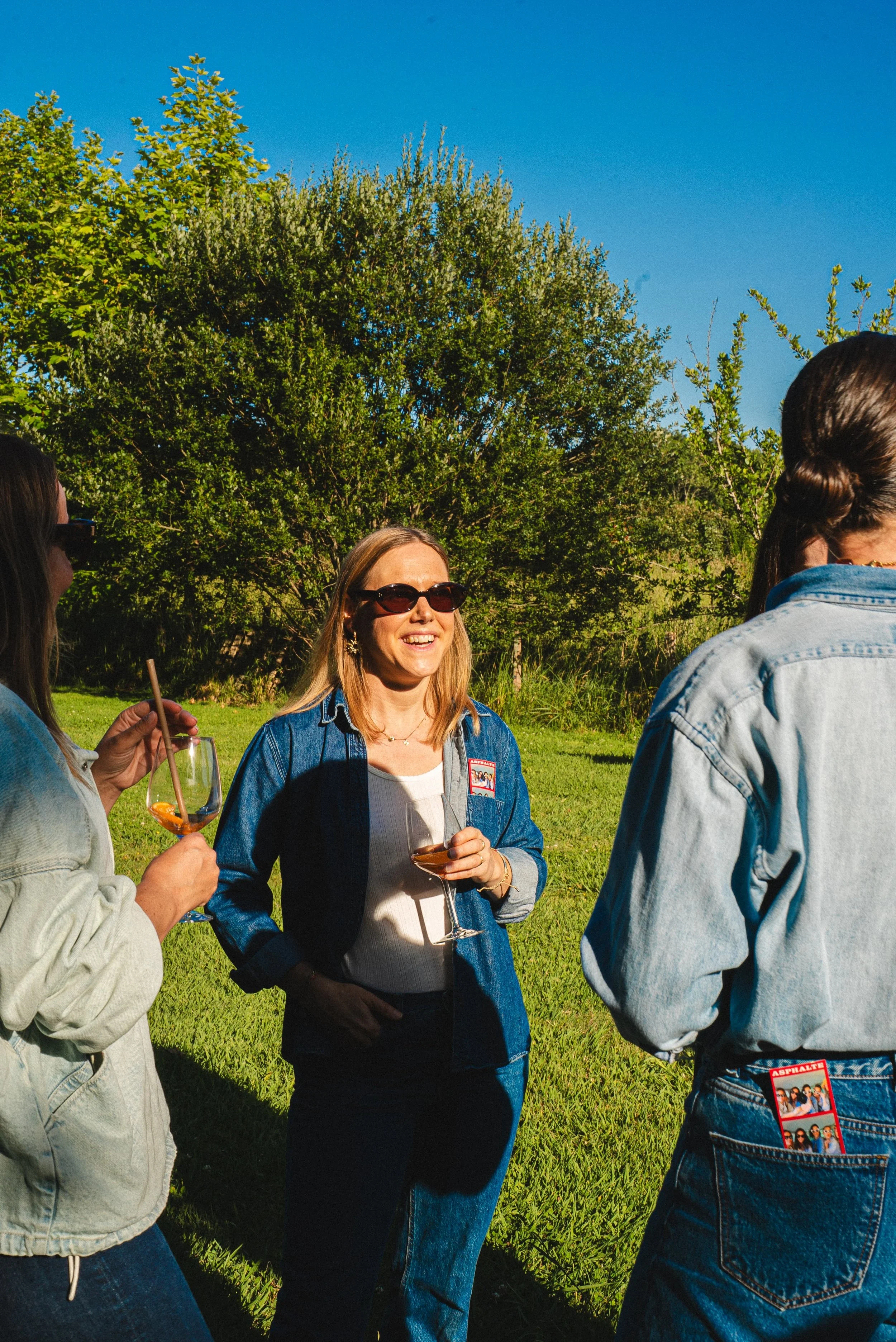 Groupe de personnes discutant dehors, une femme porte des lunettes de soleil et tient un verre de vin, soleil et arbres en arrière-plan.