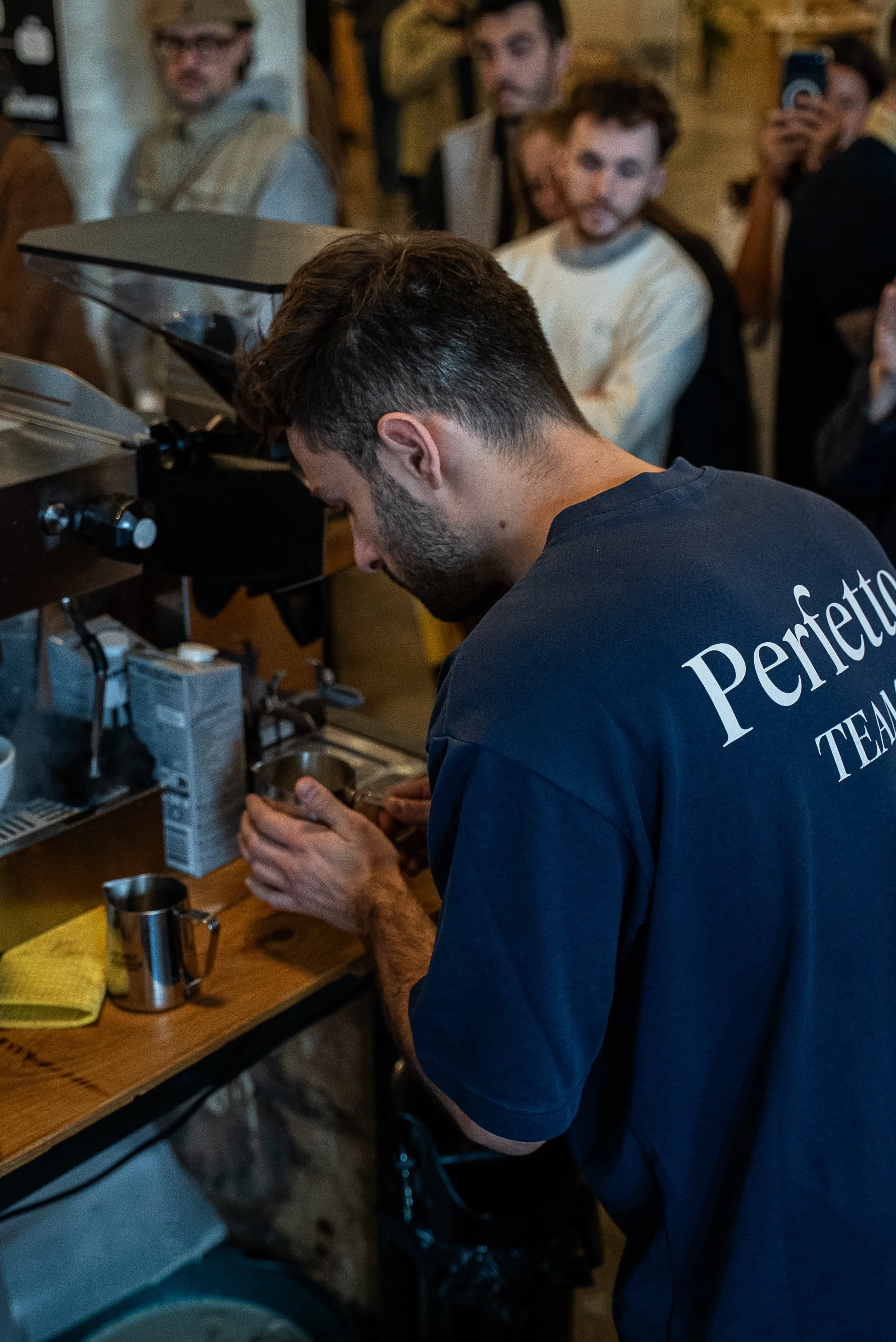 Un homme prépare une boisson derrière un comptoir dans un café ou un bar, plusieurs personnes sont présentes en arrière-plan, certaines prennent des photos ou regardent.