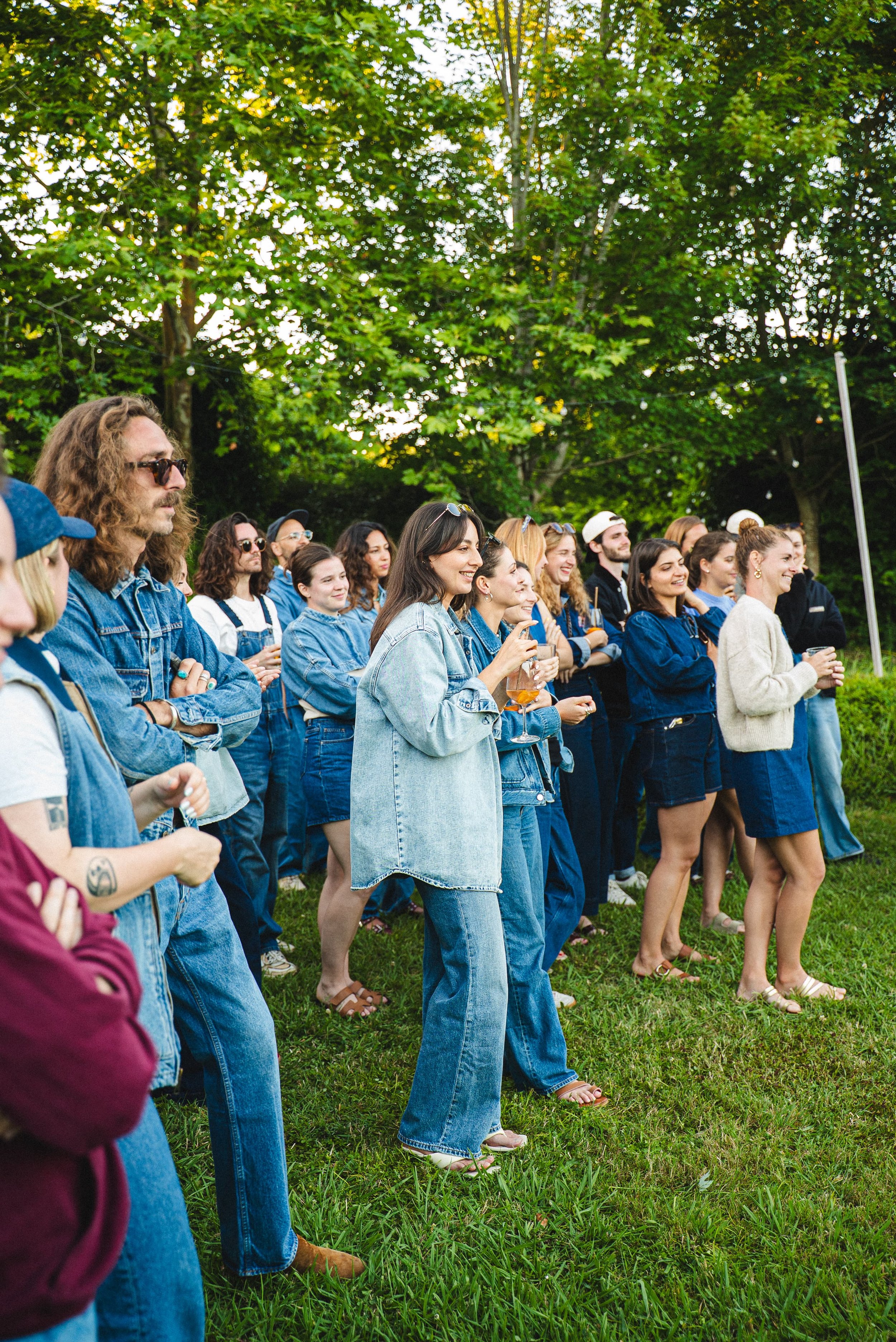 Groupe de personnes debout dans un parc, souriantes et tenant des verres, entouré d'arbres verts.