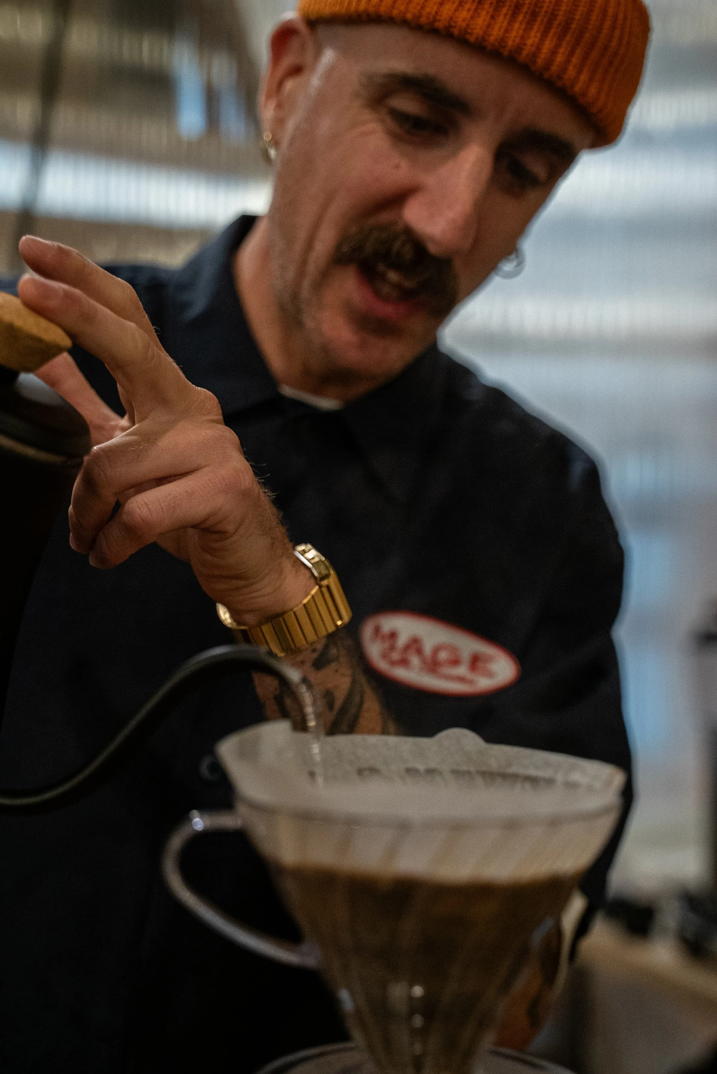 Masculin souriant portant un bonnet orange, utilisant une machine à espresso dans une cafétéria.