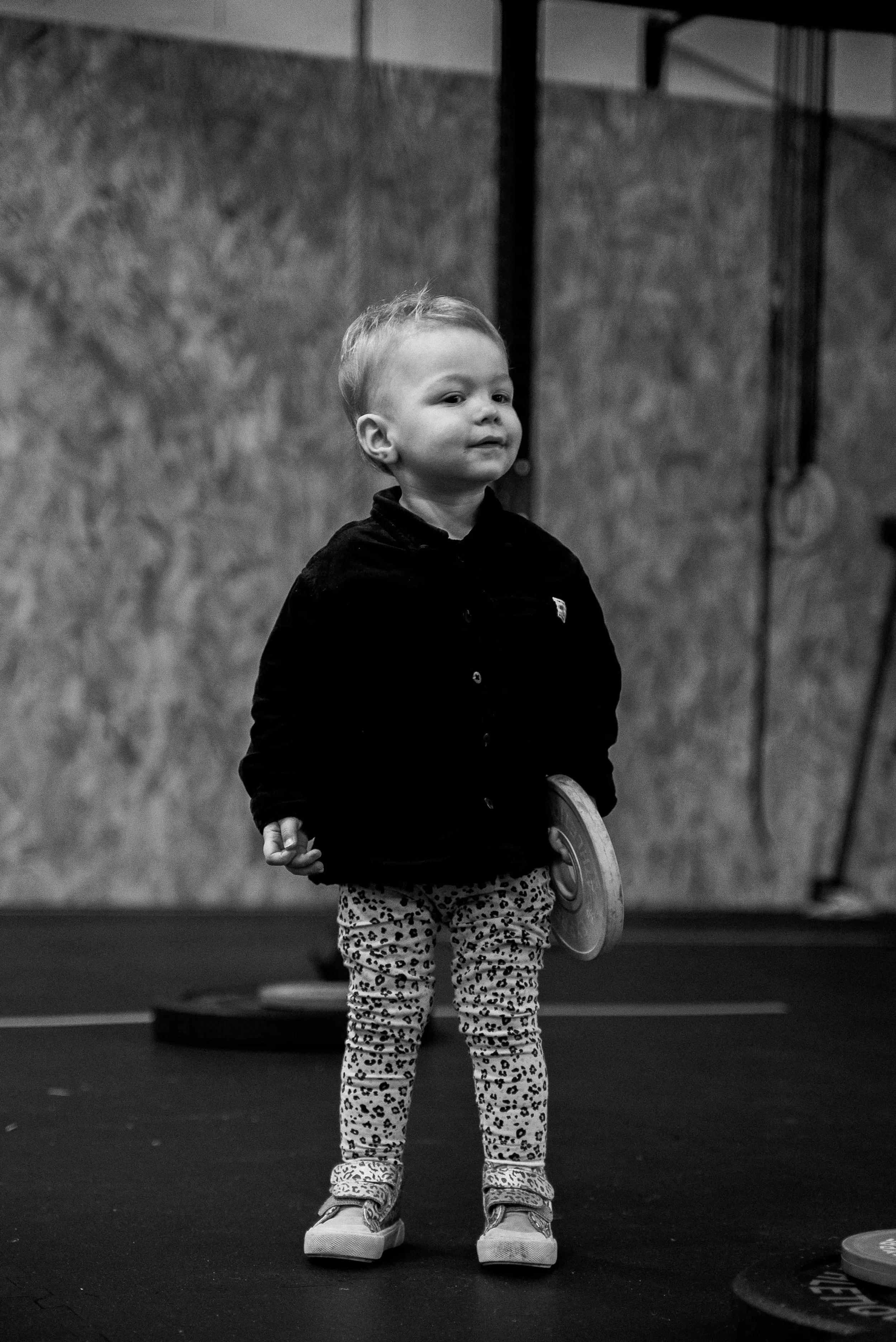 Un jeune enfant debout, portant des chaussures volumineuses, un pantalon à motif animal et un haut noir, tenant un disque en bois, dans un espace intérieur