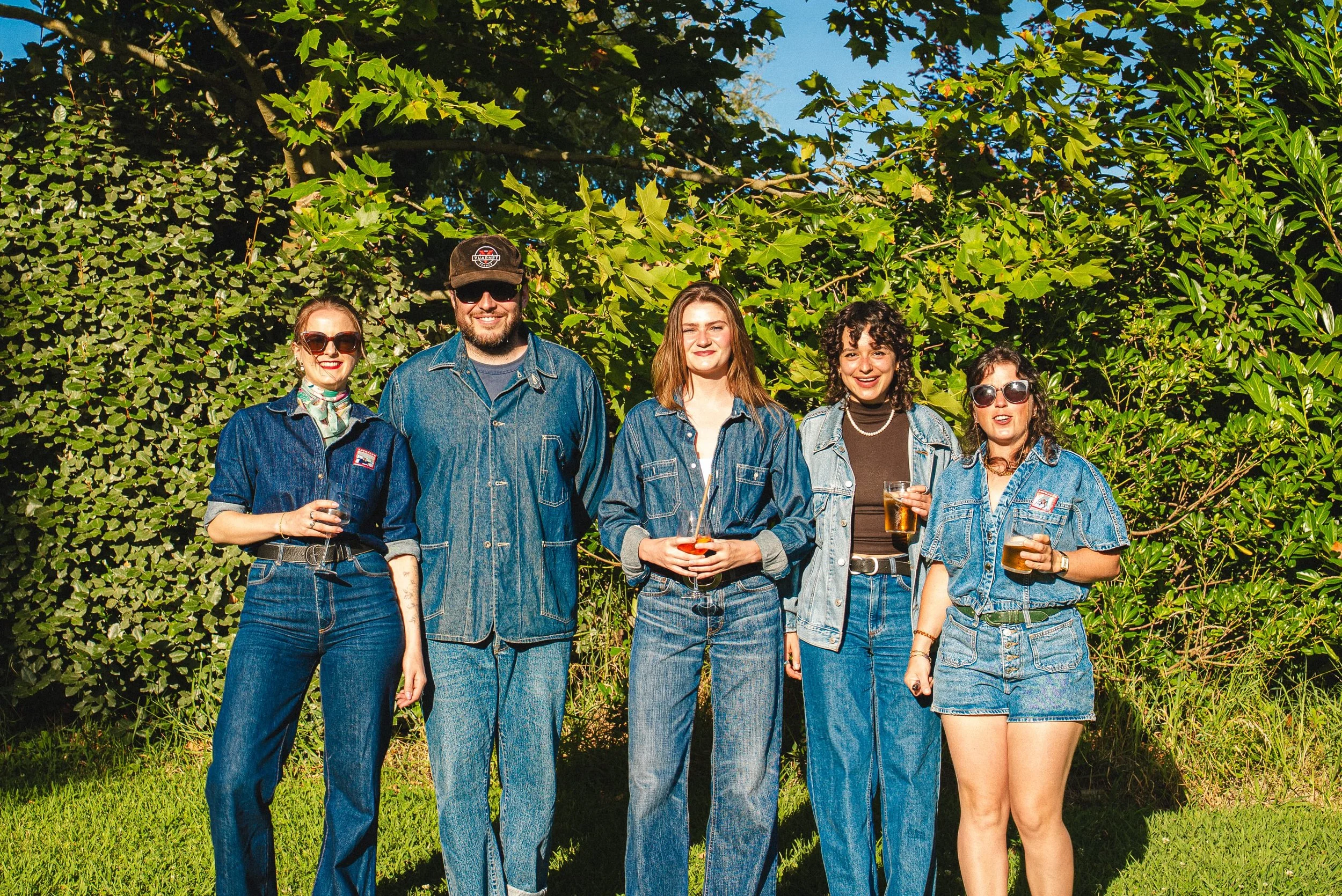 Groupe de cinq jeunes adultes debout devant un mur de feuillage dense, portant des vêtements en jean, certains avec des lunettes de soleil, tenant des verres de boissons, sous un ciel bleu ensoleillé.