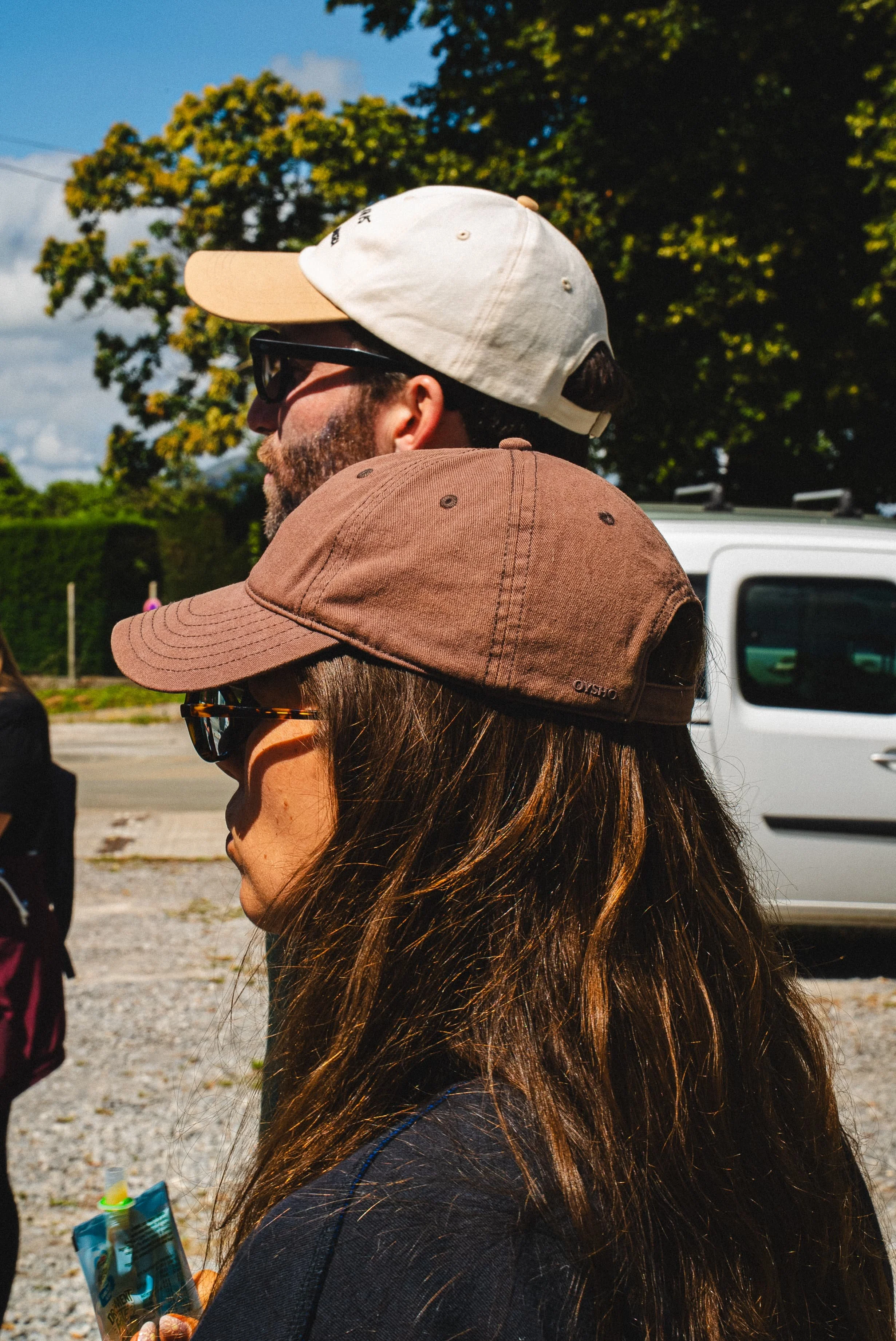 Un groupe de personnes portant des casquettes et des lunettes de soleil, vue de profil, sous un ciel partiellement nuageux, avec des arbres en arrière-plan